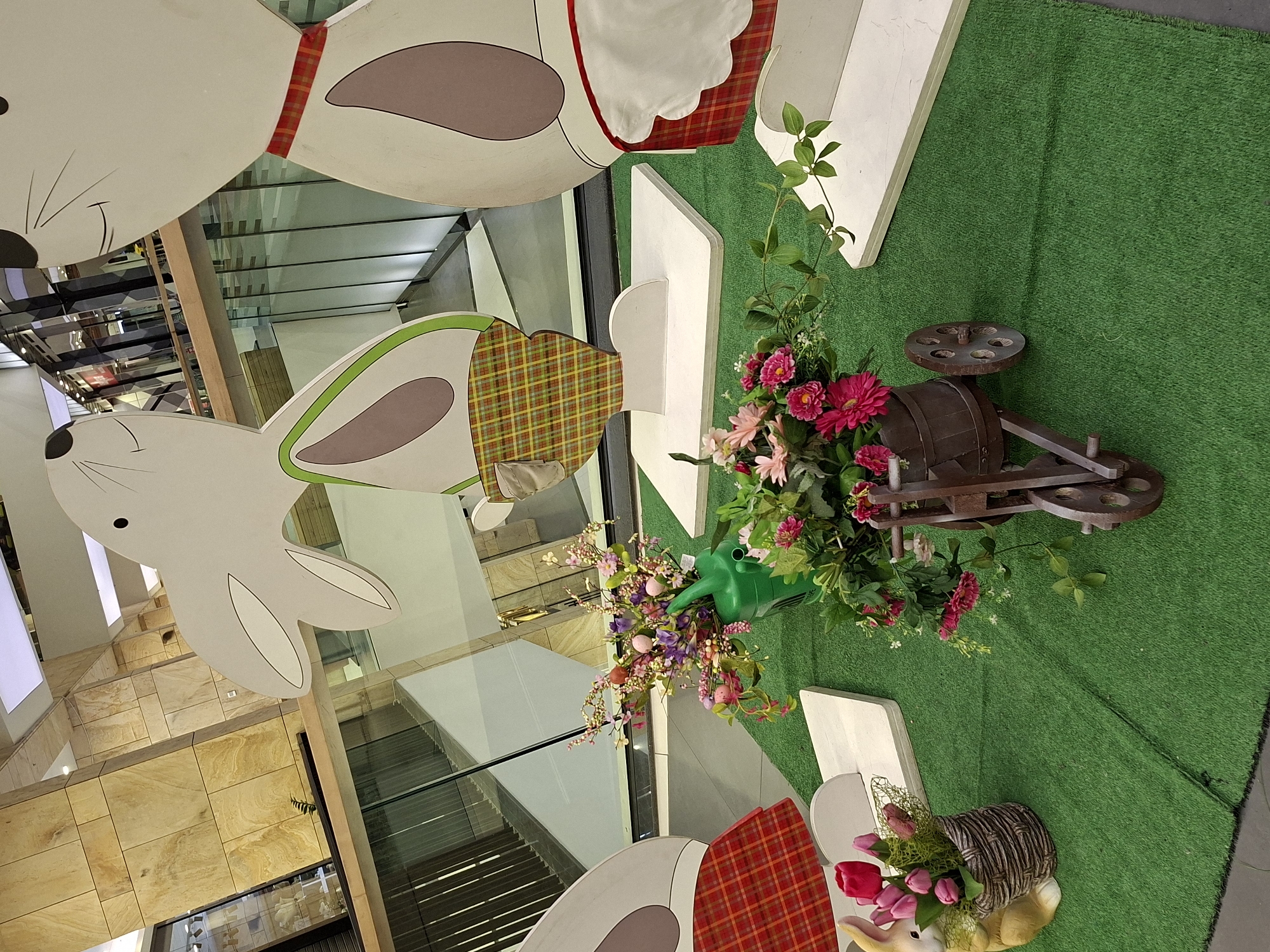 The image shows an Easter-themed decoration inside a commercial building. The decoration features large, flat bunny cutouts, some with plaid fabric accents, suspended from the ceiling. Below, on a surface covered with artificial green turf, is an arrangement including a small wooden cart filled with artificial flowers, a green watering can also filled with flowers, and a woven basket containing artificial tulips. White wooden picket fence segments are placed around the display. The building's architecture is visible through the glass above, showing a multi-level interior with stone or tile walls and metal supports. The location is likely a shopping mall or similar commercial space. The city of Plovdiv, Bulgaria is not evident within the image.