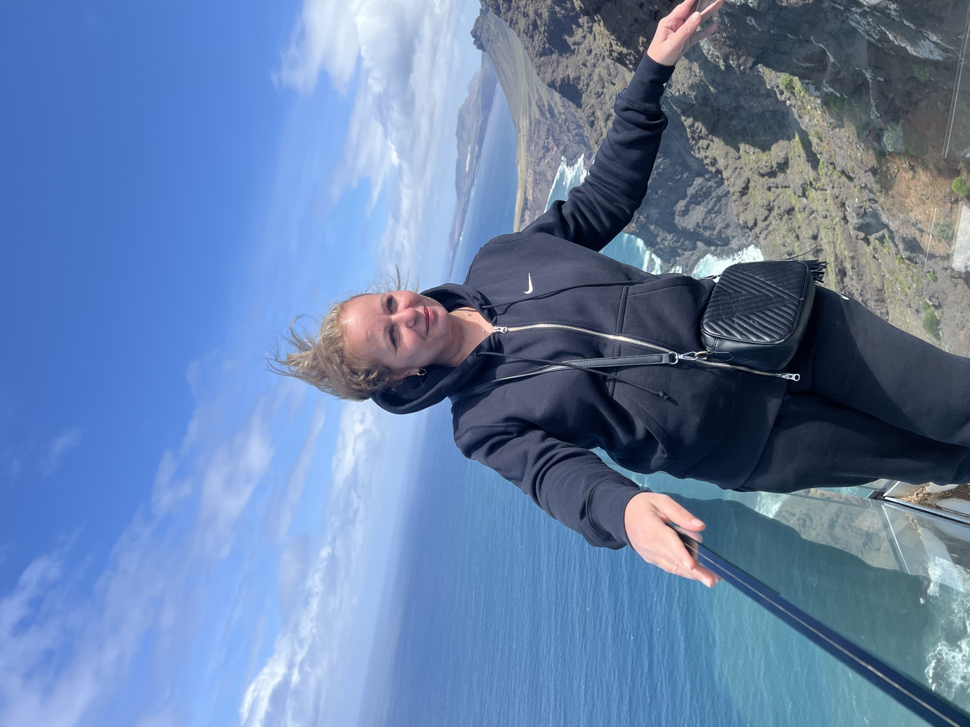 The image shows a woman standing on a glass platform overlooking a cliff and the ocean in Tirma, Spain. She is wearing a black Nike zip-up hoodie, black pants, and a black crossbody bag. She is holding a selfie stick and smiling at the camera. The sky is blue with some clouds. The cliffs are rocky and rugged. The ocean is a deep blue color.