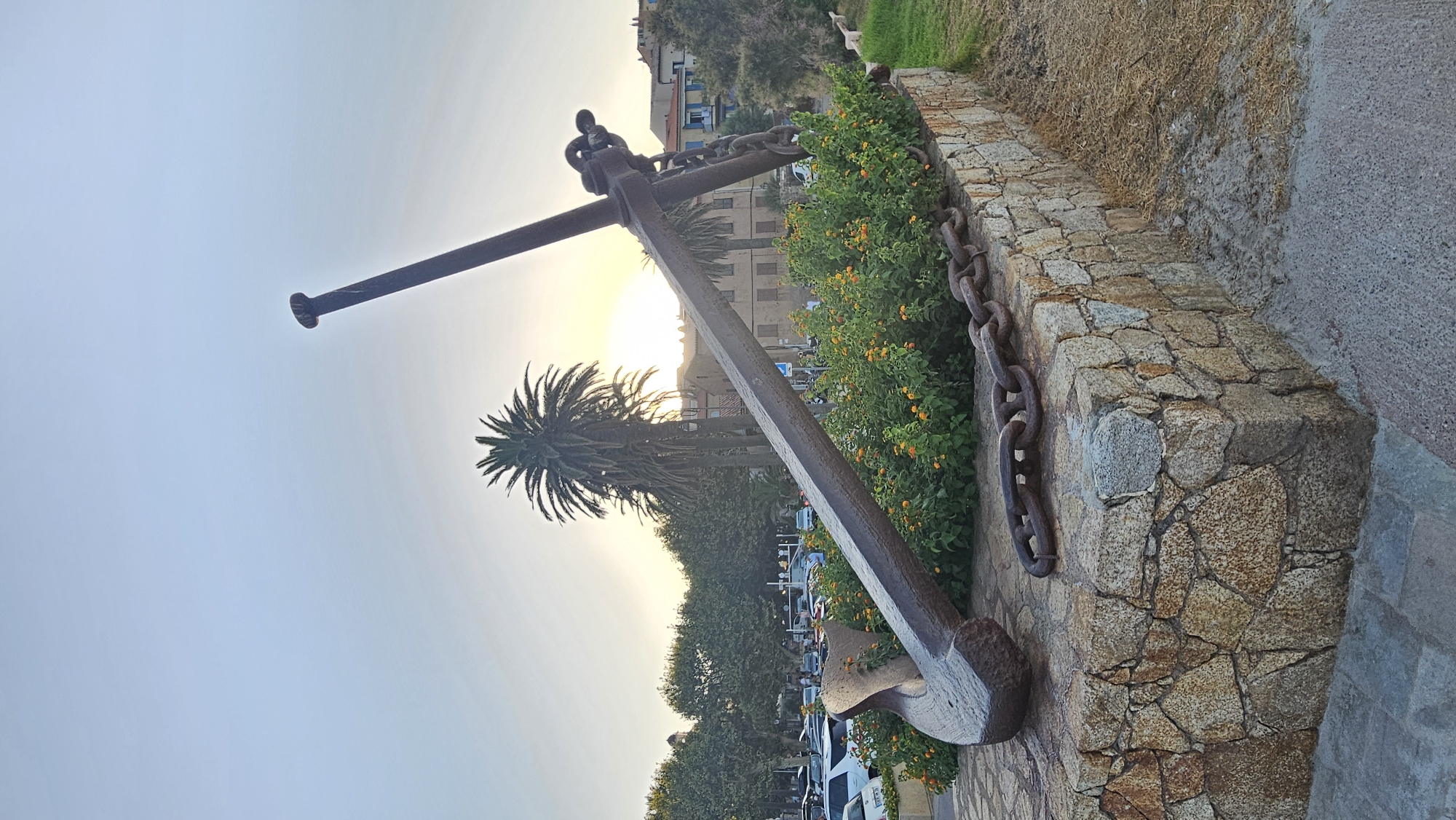 An oversized anchor and chain, likely a monument, is displayed outdoors in L'Île-Rousse, France. The anchor, made of dark metal, rests on a stone pedestal adorned with flowering bushes and a palm tree. The sun is setting, casting a warm glow over the scene and silhouetting a palm tree. In the background, buildings and parked cars suggest an urban environment near a waterfront. The sky is clear with a gradient of light blue and pale yellow from the setting sun.