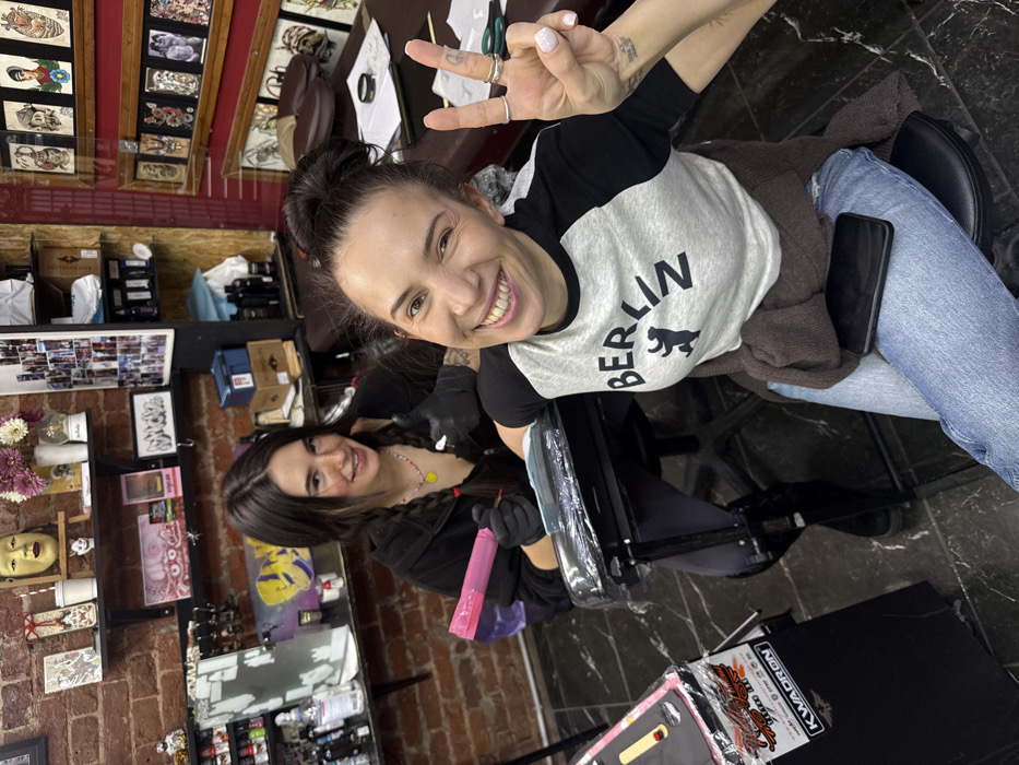 Two smiling women in a tattoo parlor. The woman in front wears a "BERLIN" t-shirt, jeans, and makes a peace sign. A brown cardigan is draped over her. Her hair is in a bun. The other woman, presumably the tattoo artist, wears black gloves and is focused on her work. The parlor has artwork on the walls, shelves with various items, and equipment for tattooing. A phone is visible on the client's lap. A tattoo supplies package sits on the lower right corner.