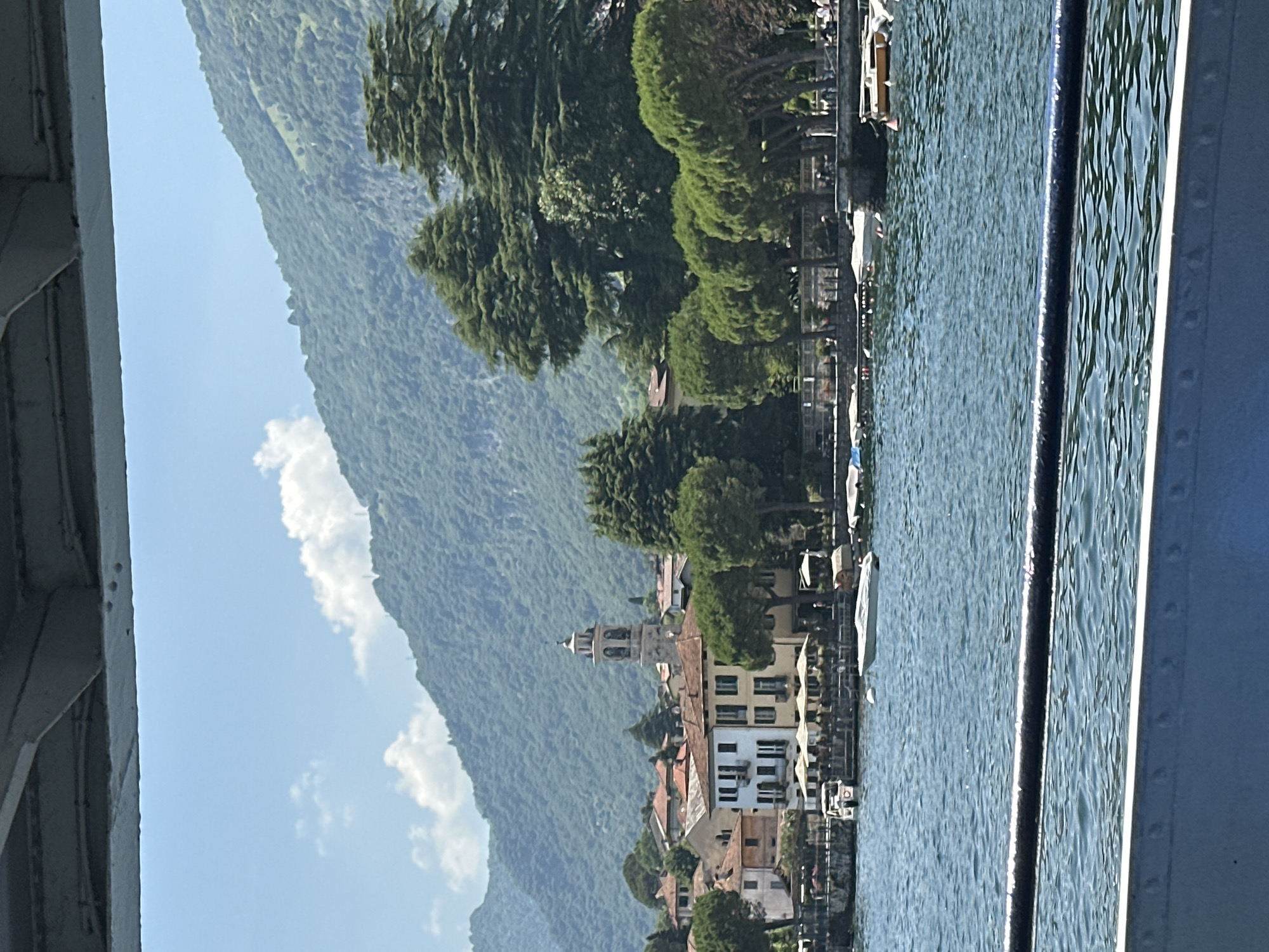 The image depicts a scenic view of the waterfront in Tremezzina, Italy, under a clear blue sky with scattered clouds. In the foreground, a dark railing and a blue metallic structure partially obscure the view of a calm body of water, likely Lake Como, which is a prominent feature of Tremezzina.
On the opposite shore, a picturesque Italian village is visible. Buildings with terracotta roofs and light-colored facades line the waterfront, interspersed with lush green trees. A tall church tower with a clock face stands out amongst the architecture, characteristic of many Italian towns. In front of the buildings, outdoor seating areas with umbrellas suggest cafes or restaurants. Several boats are moored along the shore and floating on the water, indicating a bustling aquatic environment.
Behind the village, a majestic, densely forested mountain rises, its slopes covered in vibrant green foliage, suggesting a natural backdrop. The overall impression is one of a tranquil and idyllic lakeside setting, capturing the essence of Tremezzina's charm.