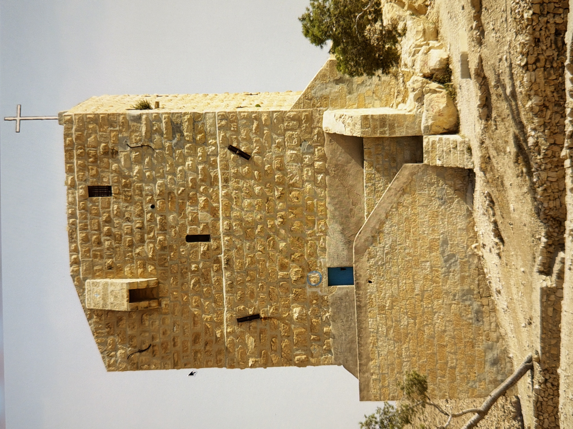 The image shows a stone building with a cross protruding from its side. The building is constructed from rough-hewn, light-colored stones. A small dark rectangular opening is visible on the left side of the building. Further to the right, a larger rectangular opening with a blue-tinted window is present. The building is situated on a rocky, uneven terrain, with a tree growing on the right side. The sky is overcast and light-colored. The overall impression is one of a solitary structure in a rugged natural setting. There is no text visible in the image.