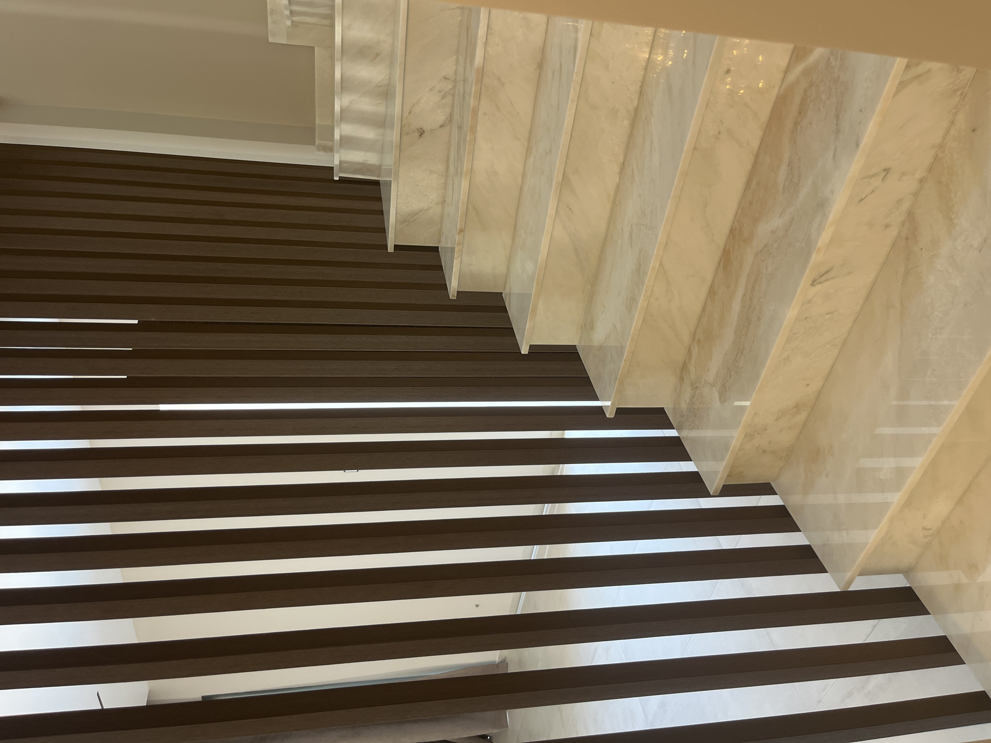 The image shows an interior view of a modern staircase with marble steps and a slatted wooden partition. The partition consists of vertical, dark brown wooden slats arranged closely together. Behind the partition, a white wall and what appears to be a window or opening with a light-colored curtain are visible. The staircase itself is made of polished marble with a creamy beige color and distinct veining patterns. The steps are wide and well-lit, reflecting the ambient light. The overall impression is one of a luxurious and contemporary interior design. The scene appears to be in a residential or commercial building located in Moni Community, Cyprus. There are no people or animals visible in the image.