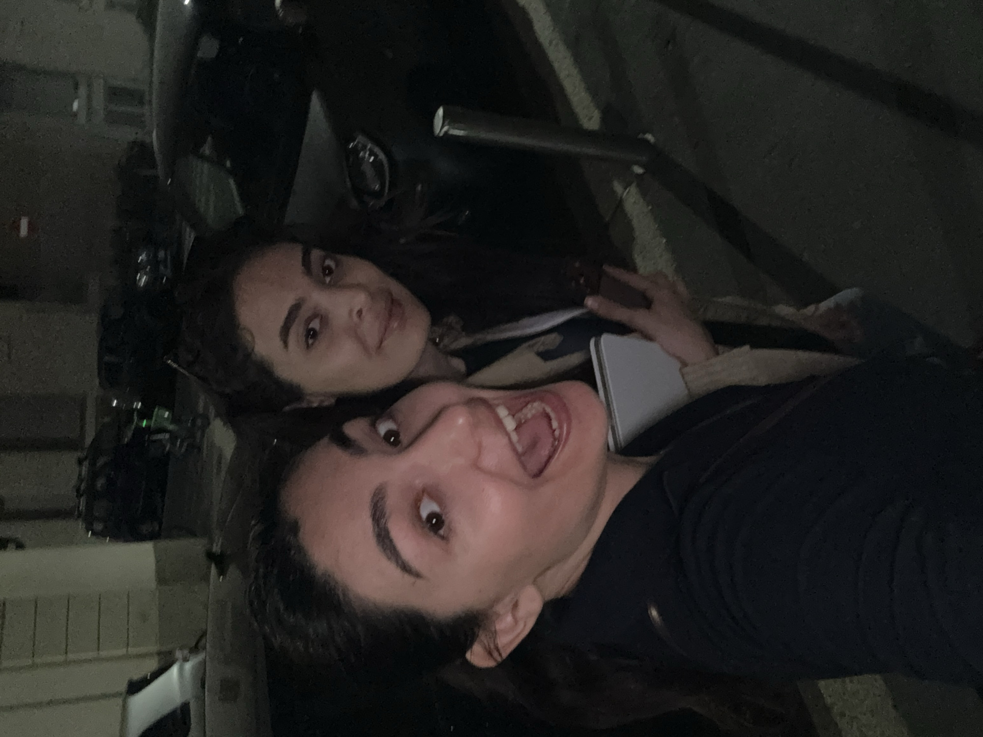 The image shows two young women taking a selfie at night. They are standing on a street with parked cars and scooters visible in the background, along with buildings across the street. The woman in the foreground is making a surprised face with her mouth open, while the woman behind her has a more neutral expression. The location is possibly in Milan, Italy. The lighting is dim. One of the women is holding a phone.