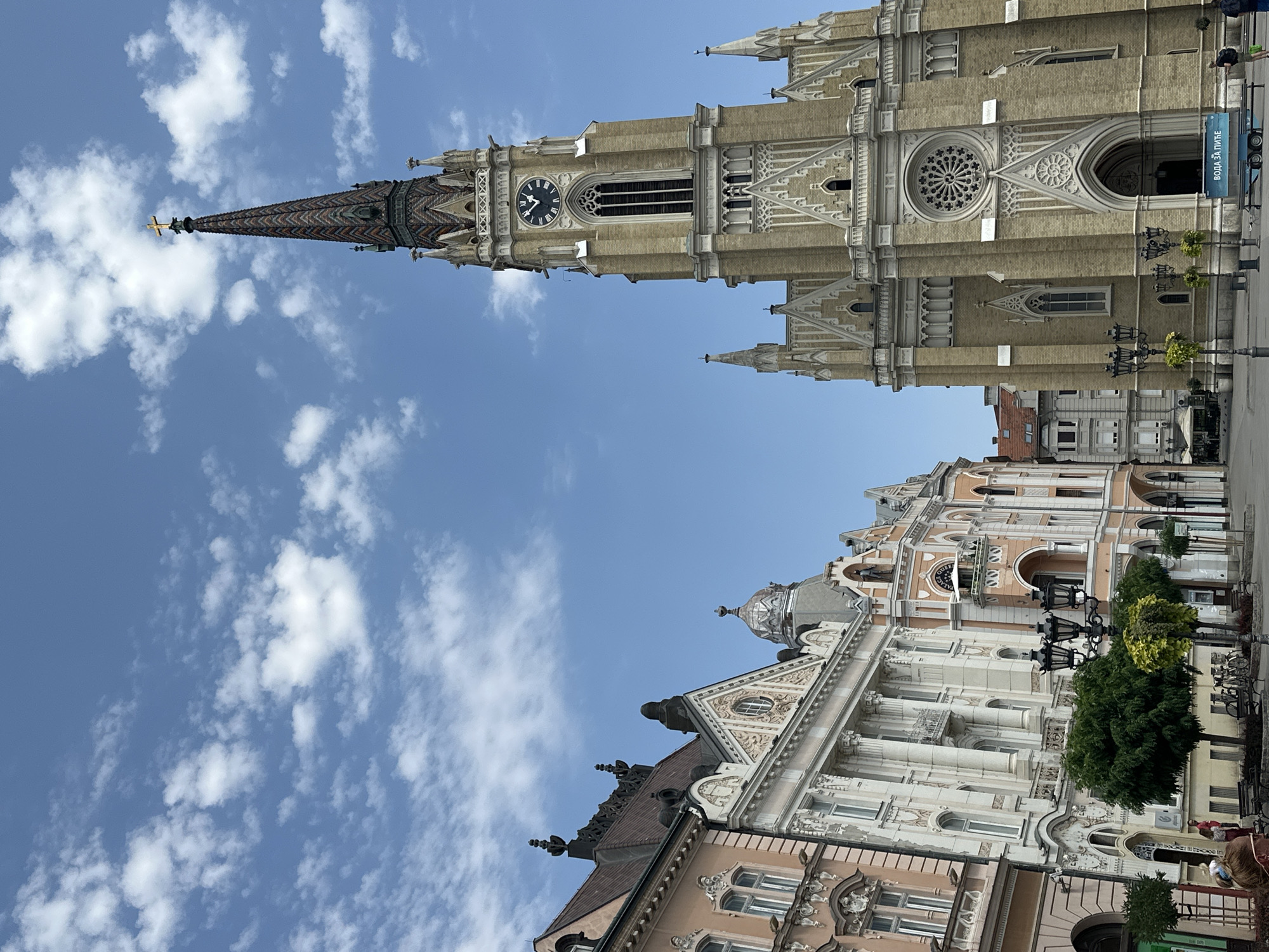 The image displays a street view of Liberty Square in Novi Sad, Serbia. On the right is the imposing Catholic Name of Mary Church, a neo-Gothic structure with a tall, ornate spire topped by a golden cross. The church features a large rose window above its arched entrance, and its facade is adorned with Gothic architectural details. To the left of the church are several historic buildings with classical and Art Nouveau influences. These buildings have colorful facades, decorative stonework, and elaborate balconies. The sky above is a bright blue with scattered white clouds. In the foreground, there are street lamps, trees, benches, and a few people can be seen walking and standing in the square. A blue trailer with Cyrillic text, reading "ВОДА ЗА ПИТЬЕ" (Drinking Water), is parked in front of the church. The overall scene suggests a sunny day in a bustling European city square.