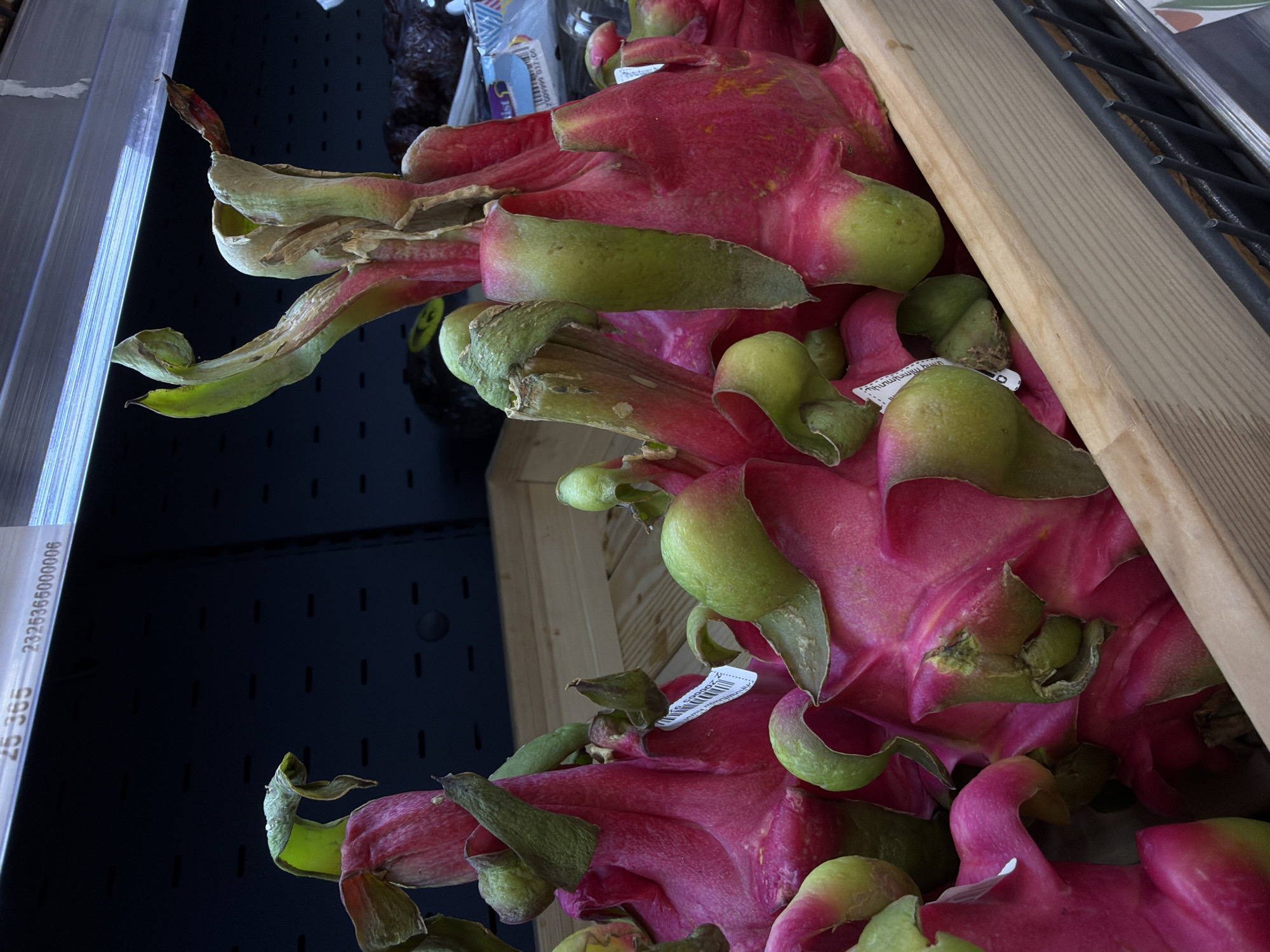 The image shows a close-up of dragon fruits in a crate, likely at a market or grocery store. The fruits are characterized by their pink skin and green, scale-like leaves. A wooden plank of the crate is visible to the right. The scene is possibly located in Yerevan, Armenia, considering the query's specification. The image lacks any people or interactions.