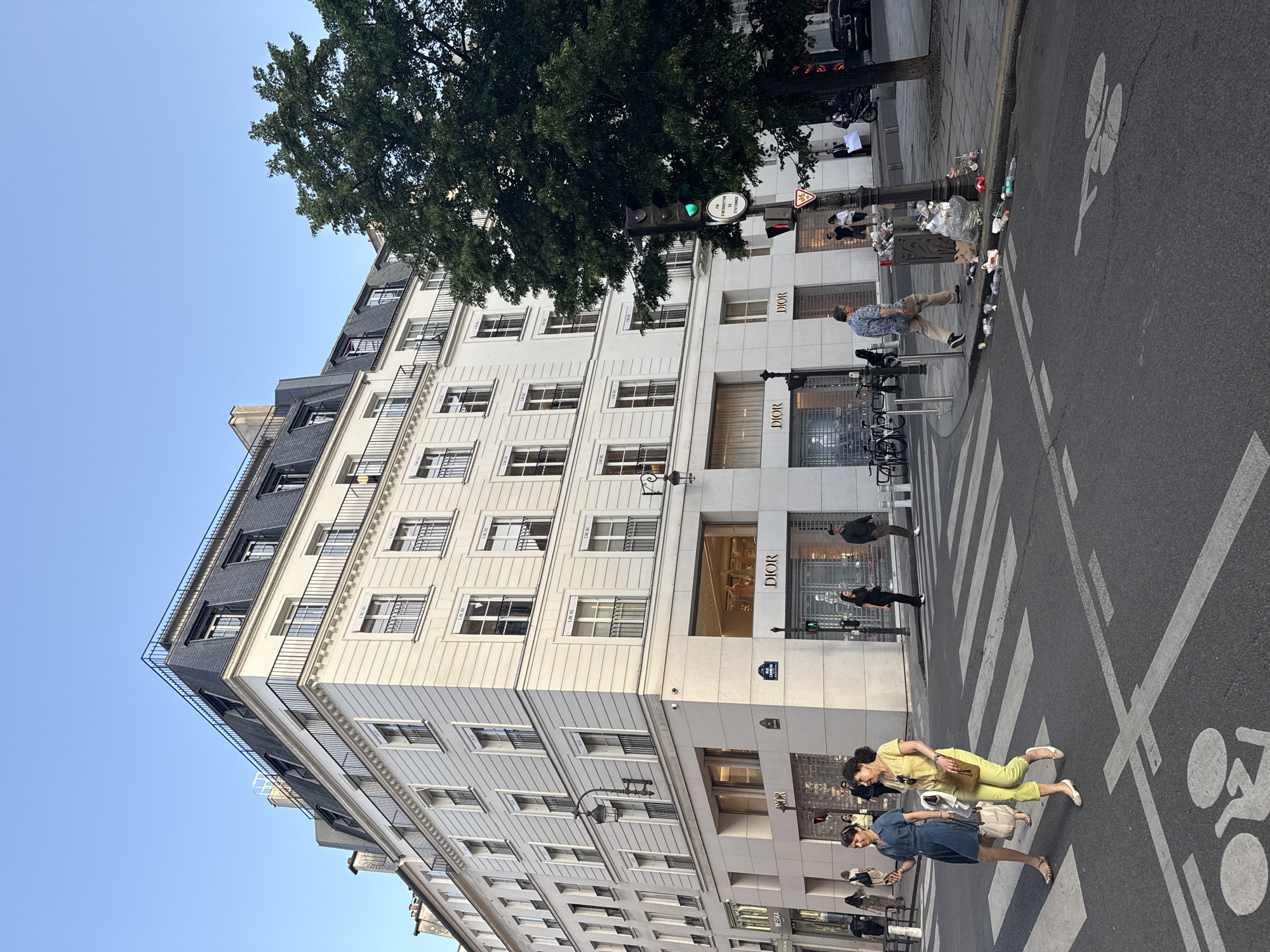 The image captures a street scene in Paris, France, featuring a building prominently displaying the "DIOR" brand logo on its façade. The building is multi-storied, with numerous windows and a light-colored exterior. A large tree is situated near the building, partially obscuring its upper floors. Several people are walking across a pedestrian crosswalk in the foreground. A bicycle lane is marked on the street. A traffic light is visible near the Dior store, showing a green light. A street sign is mounted on the corner of the building.