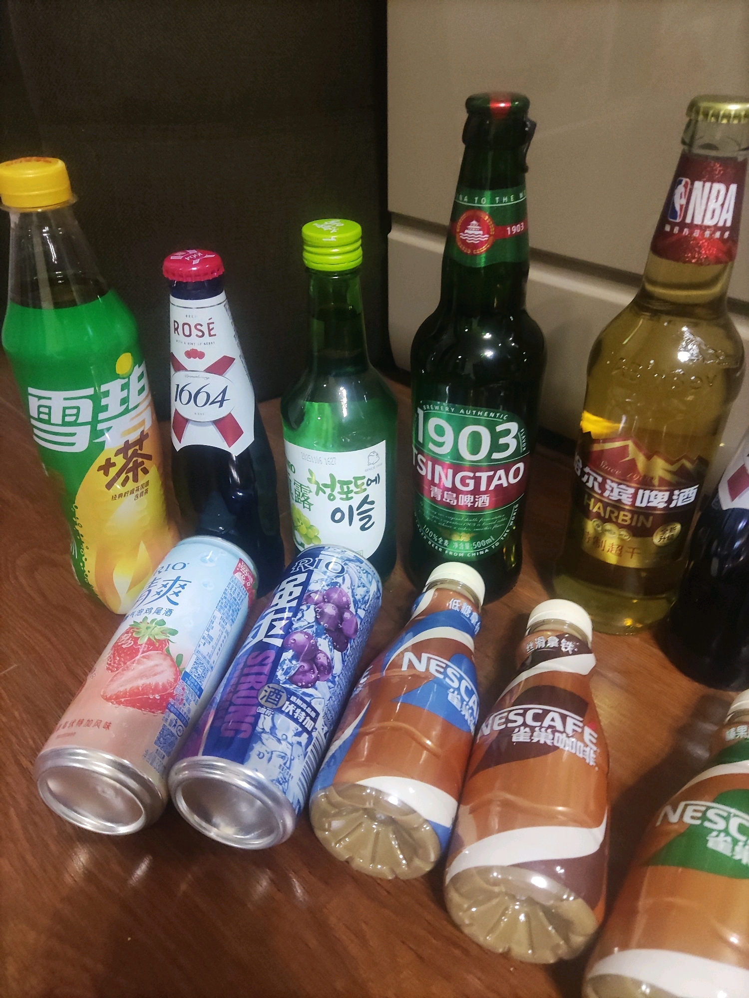 The image displays a collection of assorted beverages arranged on a wooden surface, possibly a table or floor, against a dark, textured background. The items include several bottles and cans of various drinks.
On the left, a green bottle with a yellow cap contains a light-colored liquid, possibly a soft drink or juice, with Chinese characters on its label. Next to it is a white bottle with red accents and the text "ROSE 1664". A green bottle with a green cap and white text, which appears to be a Korean beverage, is positioned further to the right.
In the foreground, two aluminum cans are visible. The left can has an image of strawberries and text in what appears to be Chinese. The right can features an image of grapes and also has Chinese text.
Several bottles with dark green caps and labels are arranged to the right. One green bottle has the text "1903 TSINGTAO" and Chinese characters. Adjacent to it is a yellow-tinted bottle with "HARBIN" in English and Chinese characters. Another dark-colored bottle is partially visible on the far right.
Three plastic bottles with brown and white labels, featuring the "NESCAFE" logo and Chinese text, are arranged in the lower right portion of the image. These appear to be coffee drinks.
The lighting in the image suggests it was taken indoors, possibly in a dimly lit room. The wooden surface has a visible grain pattern and reflects some light.
This scene does not provide any information that would allow for its location to be identified as Artyom, Russia, or any other specific city. The objects and arrangement are typical of a casual gathering or personal collection of beverages.