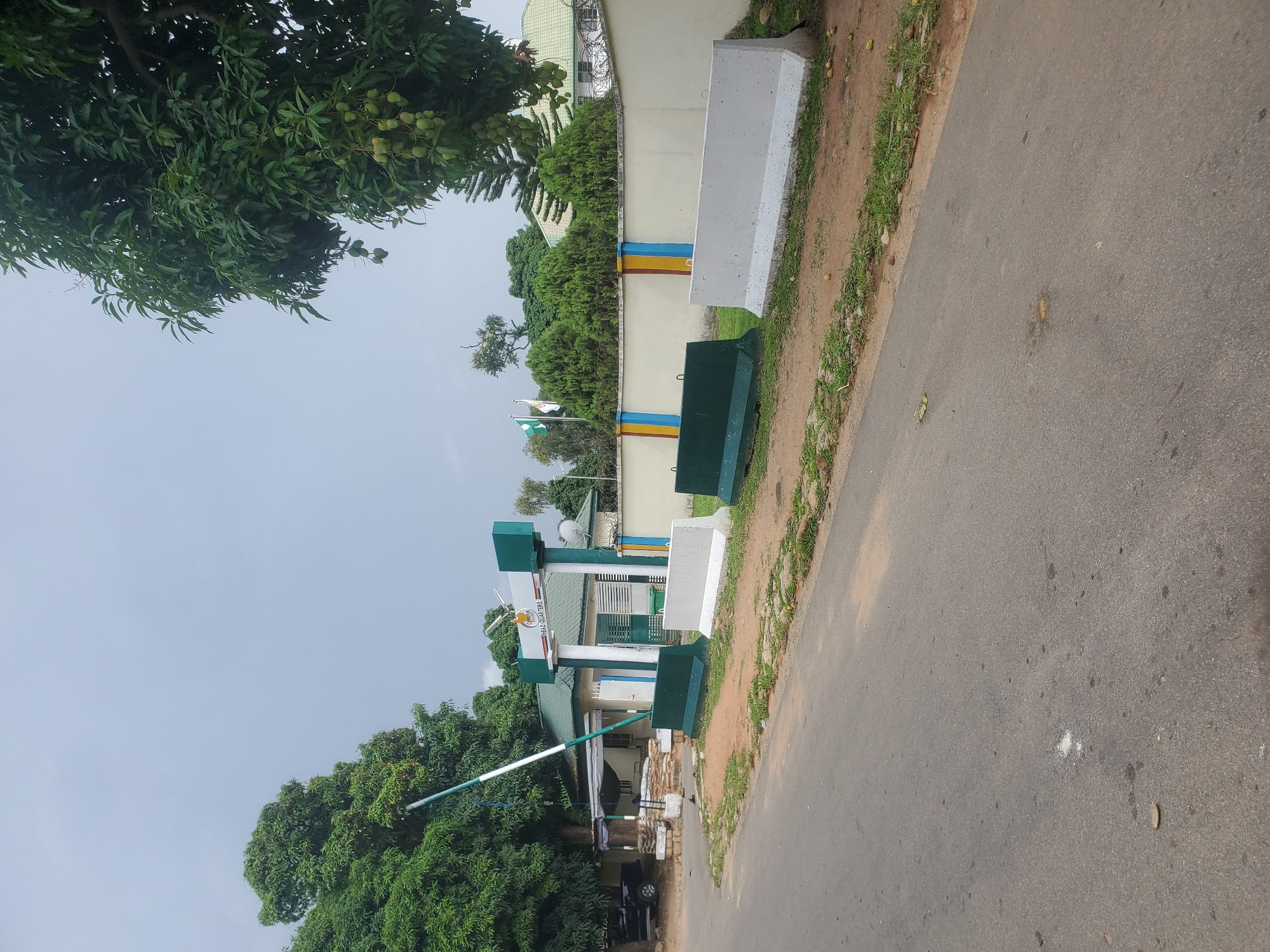 The image captures the entrance to a building in Jos, Nigeria, identified by a sign as "HEAD QUARTERS". The building has a green roof and is surrounded by a light-colored wall topped with barbed wire. In front of the building, a retractable barrier is partially raised, indicating it is either opening or closing. Several concrete barriers, some painted dark green and others white, are placed along the roadside, suggesting a security checkpoint or traffic management. Trees, some bearing fruit that appears to be mangoes, are visible on either side of the building and in the background. The sky is overcast. The road in the foreground is paved and shows signs of wear and tear, with some scattered debris and vegetation along the edges. A black vehicle is parked to the left of the building, partially obscured by a tree. There are no people clearly visible in the image.