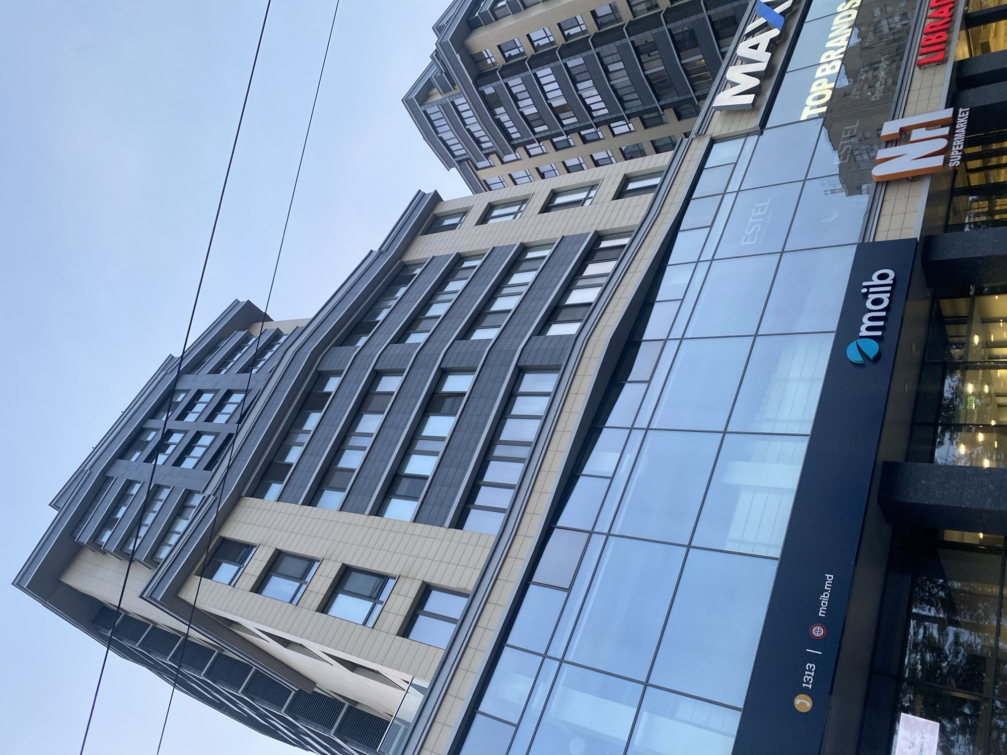 The image shows a modern, multi-story commercial and residential building with a glass facade on the lower floors. Several businesses are visible on the ground floor, including "maib," "MAXI," "TOP BRANDS," "N-1 SUPERMARKET," and "LIBRARI." The "maib" logo is a stylized turquoise "m" with a teal oval. The "MAXI" logo features the word in white with a blue "V" shape. "TOP BRANDS" is written in white capital letters. "N-1 SUPERMARKET" is displayed on an orange and white sign. The building is located in Chișinău, Moldova, and appears to be in a commercial area. The sky is overcast, and power lines are visible crossing the frame. The architecture is contemporary, with a combination of beige and dark gray building materials. The perspective is from a low angle, looking up at the building.