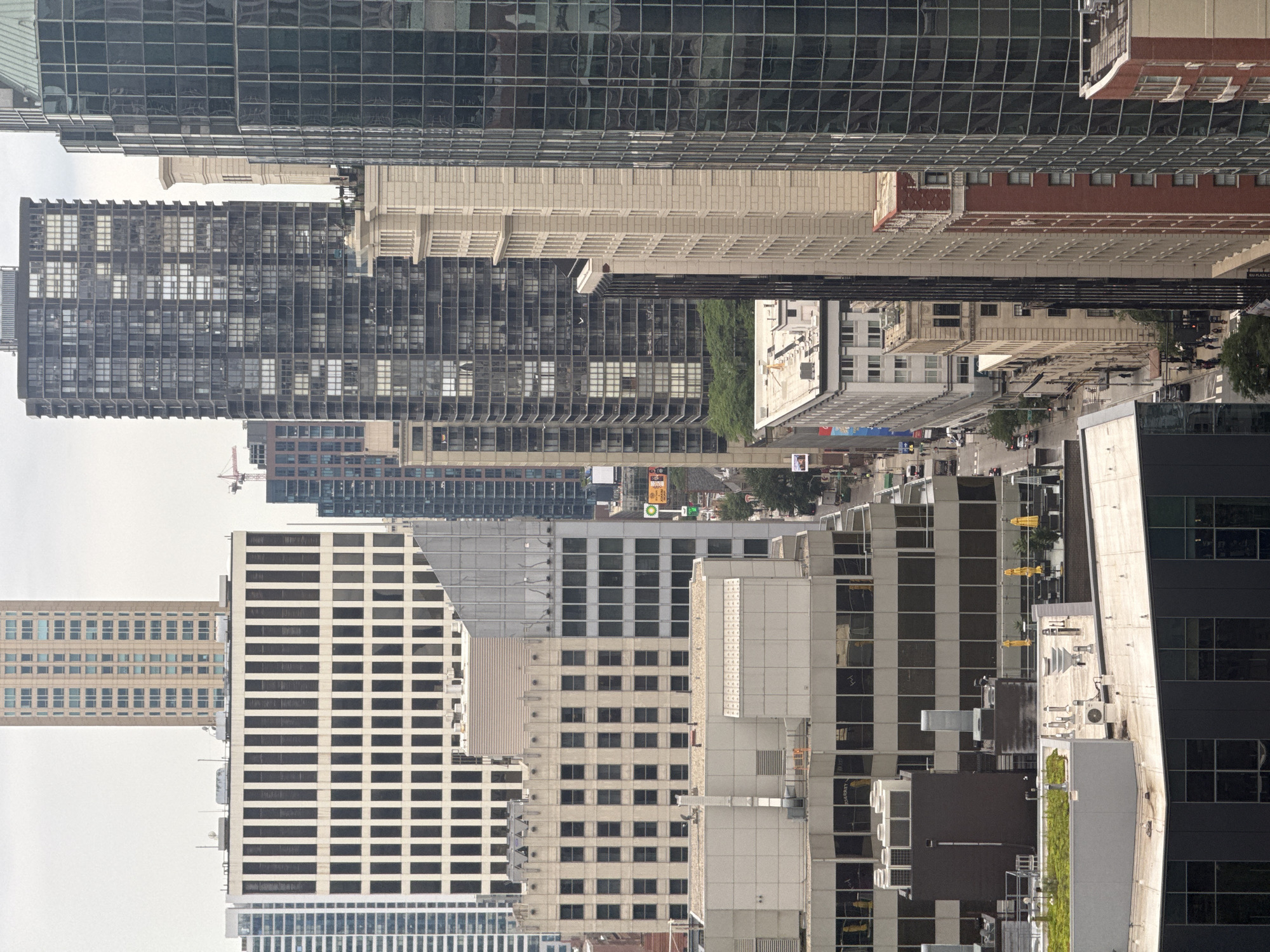 This is an aerial, vertical-orientation photograph of a dense urban landscape, likely taken in Chicago, United States of America. The scene is dominated by a collection of modern and older skyscrapers, showcasing a variety of architectural styles.
The most prominent subjects are the buildings themselves, with their distinct facades, window patterns, and heights. We see sleek, glass-fronted buildings with grid-like reflections, contrasted with older structures featuring brickwork and more traditional window arrangements. Some buildings have visible air conditioning units and rooftop structures, suggesting the image was taken on a clear day with good visibility.
Below the towering buildings, a street scene is visible. Cars and what appear to be people are present, indicating ongoing activity. The presence of traffic lights, street signs, and the general layout of the street suggest a bustling urban environment.
No specific event or activity is clearly depicted beyond the everyday life of a city. The lighting suggests it is daytime, possibly late morning or early afternoon, with the sun casting some shadows but not creating extremely harsh contrasts. The overall impression is one of a vibrant, active city from a bird's-eye view.
There is some visible text on a sign in the distance, appearing to be a street sign or advertisement. The text is too small to be legible.