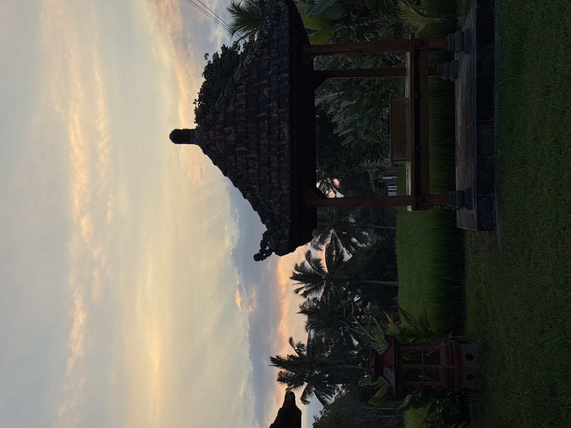 This is a photograph taken in Tegallalang, Indonesia, at sunset. The image shows a traditional Balinese gazebo in the foreground, constructed of dark wood with a tiled roof. The gazebo is situated on a stone platform, surrounded by lush green grass. To the left of the gazebo, there is a red decorative lantern. In the background, vibrant green rice paddies stretch out, bordered by a dense line of palm trees and other tropical foliage. The sky above is painted with soft hues of orange, yellow, and pink, with streaks of light breaking through the clouds, indicative of the setting sun. The overall scene evokes a tranquil and serene atmosphere typical of rural Bali.