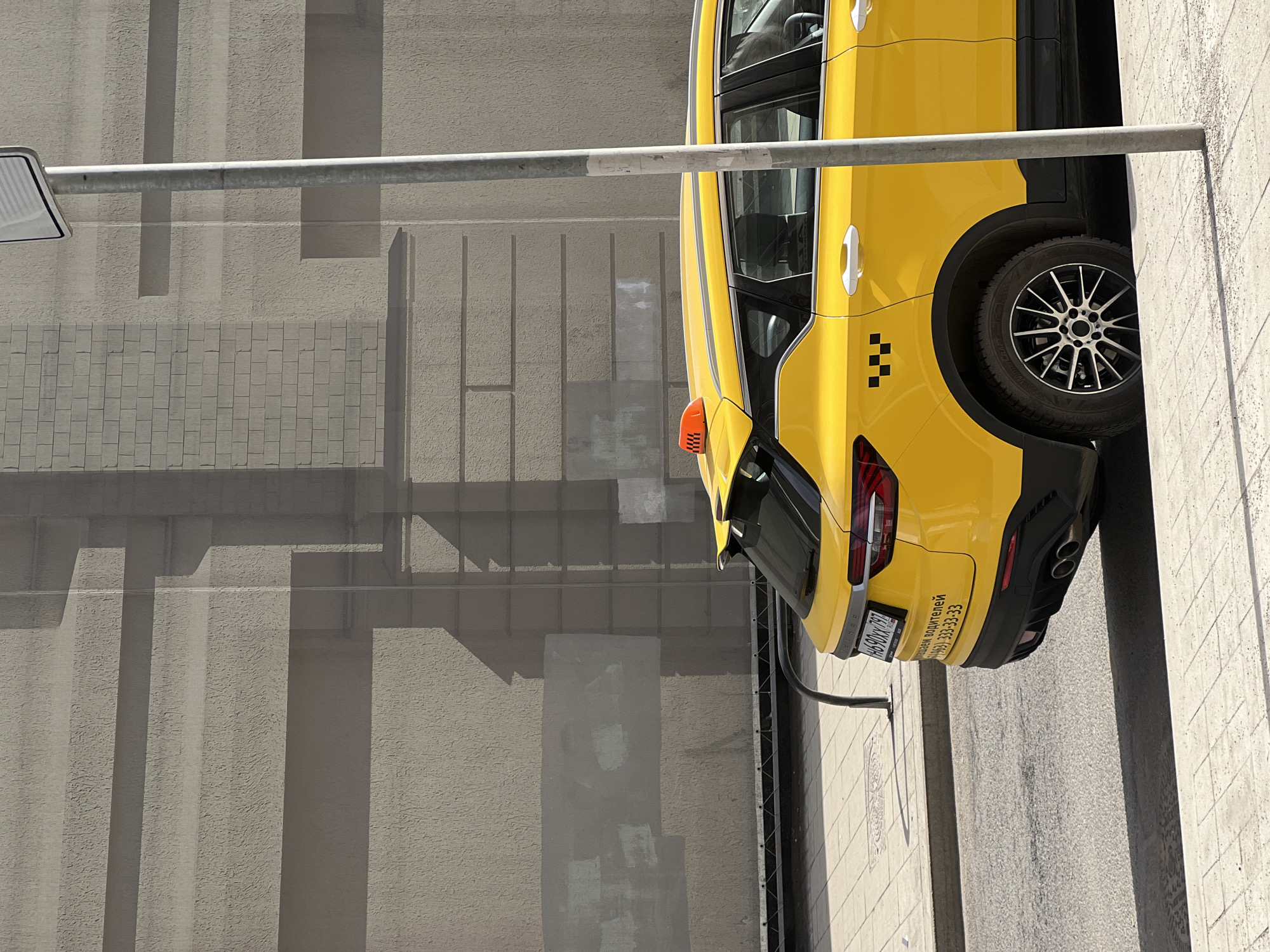 A yellow taxi vehicle is observed in an urban setting. The license plate reads "H090XY797 HA", indicating the Russian Federation. The text "телефон водителей +7(499) 333-33-33" is printed below the license plate. The vehicle is adjacent to a light grey building with vertical striations and rectangular indentations. A metal bar runs horizontally across the upper portion of the image. The location is likely Moscow, Russia, based on the license plate series (797 is frequently associated with Moscow).