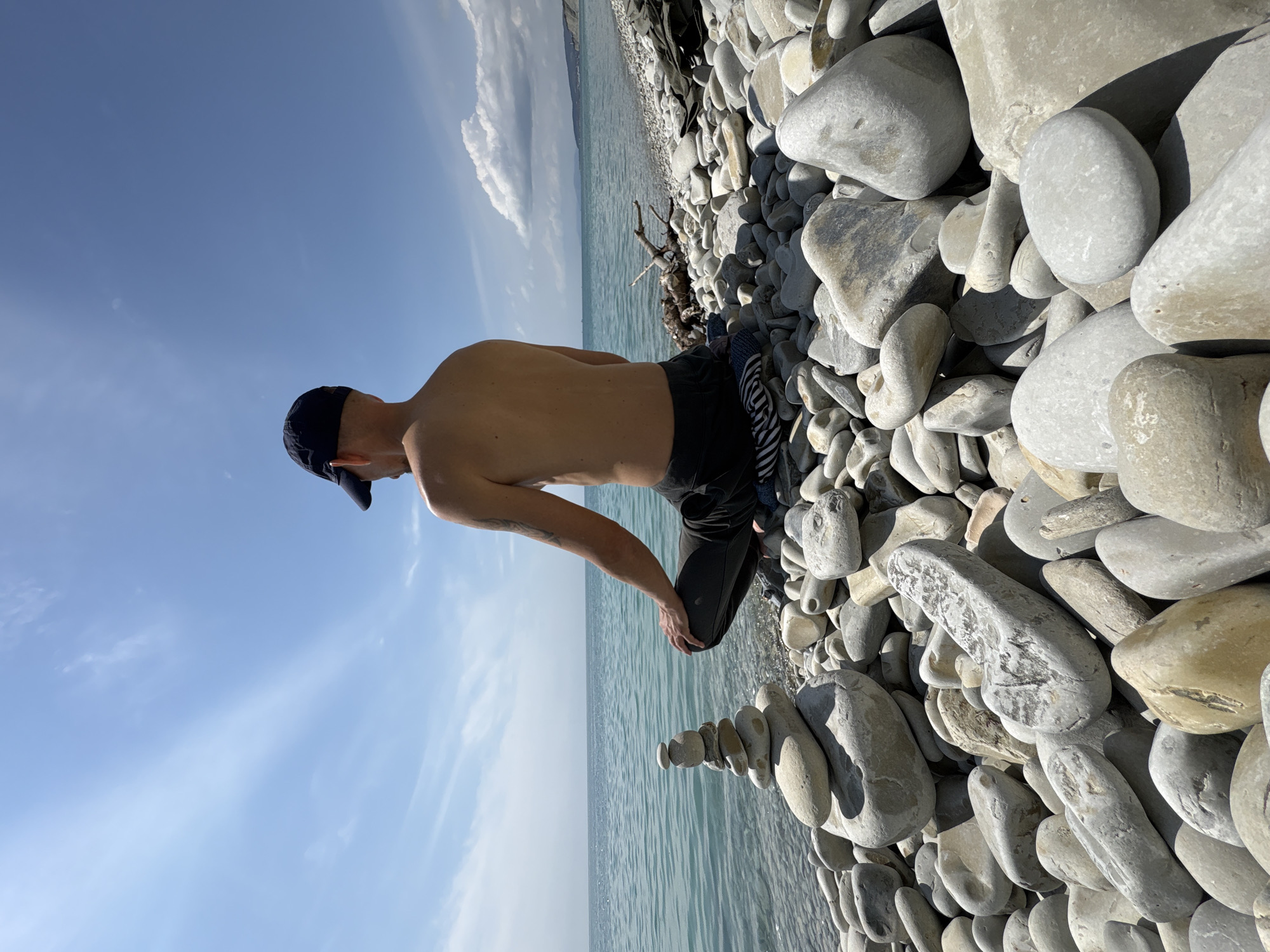 A medium shot shows a shirtless man in a black baseball cap and black shorts kneeling on a pebble beach. He is balancing a flat stone on top of a stack of stones already balanced precariously by the water's edge. The beach is comprised of smooth, grey pebbles of varying sizes. The water is a light blue color, and the sky is clear with a few scattered clouds. The location is likely a beach in Gelendzhik, Russia, given the user's specification.