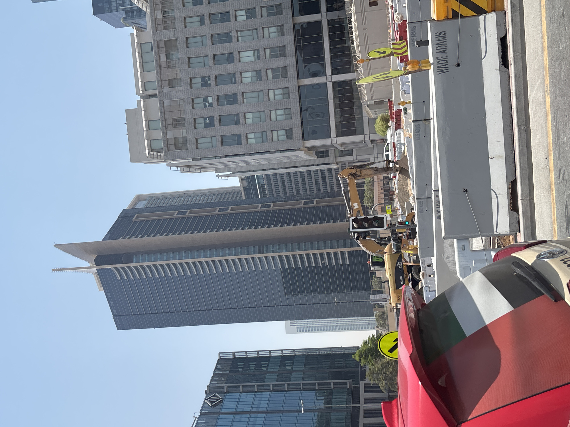 The image shows a street scene in Dubai, United Arab Emirates, with several modern skyscrapers under a clear blue sky. In the foreground, the rear of a red car with a United Arab Emirates flag decal on its window is visible. Construction barriers, some bearing the name "WADE ADAMS," line the road. Behind the barriers, a yellow excavator is operating, suggesting ongoing construction work. To the left, a glass-fronted building displays a diamond-shaped logo. Further in the background, a very tall, dark-colored skyscraper with a distinctive angled top dominates the skyline. Other buildings of varying heights and architectural styles are also present. Traffic lights and street signs are visible. The overall impression is a bustling urban environment with significant development.