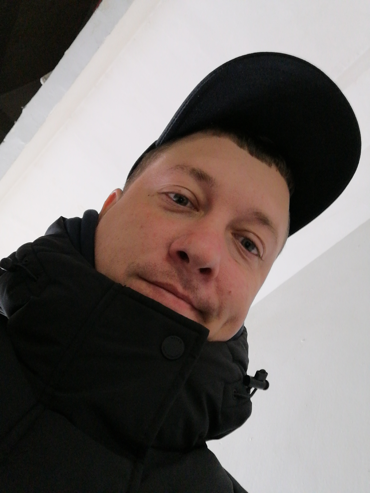 A close-up, low-angle portrait shows a man looking directly at the camera. He is a white male adult, wearing a black baseball cap and a black, possibly padded, jacket or coat with a high collar. His hair is short and visible beneath the cap, and his eyes are light-colored. He has a neutral expression, possibly with a slight smirk.
Details of his attire include a black button closure visible on the right side of his jacket collar and a black drawstring toggle on the left side, near his neck. The background is mostly plain white, suggesting an indoor setting with a wall or ceiling. A dark, indistinct area is visible in the upper left corner, possibly a shadow or part of the building's structure. The lighting appears even. No specific time of day, weather, or outdoor location cues are discernible. There is no visible text in the image.