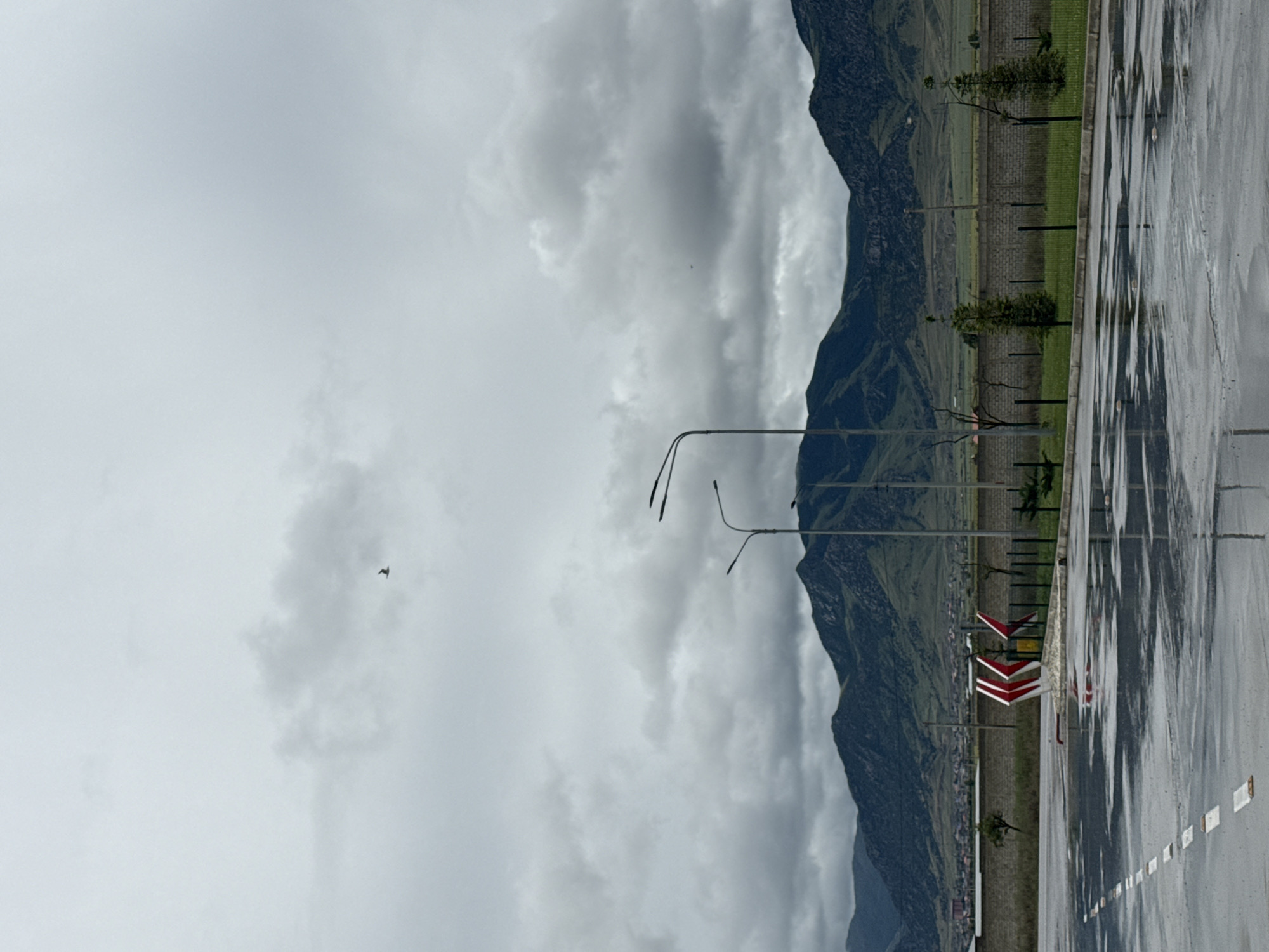 This outdoor scene, taken in Korkmaskala, Russia, captures a vast, overcast landscape with evidence of recent rain. The image is composed of a foreground featuring a wet paved surface, a mid-ground with various structures and greenery, and a background dominated by mountains under a cloudy sky.
In the foreground, a wide, wet asphalt or concrete area, possibly a parking lot or road, is visible, reflecting the grey sky and creating shimmering patterns. White dashed lines are painted on this surface.
The mid-ground includes several tall streetlights or lampposts with multi-arm fixtures. To the right, prominent red and white chevron signs indicate a curve or hazard. A strip of green grass and a low brick wall run parallel to the paved area, behind which a few small trees or bushes are scattered. Faint outlines of buildings or a small settlement can be seen at the base of the mountains in the distance.
The background is dominated by large, imposing mountains, covered in green vegetation, stretching across the entire frame. The sky above is completely overcast with thick, grey clouds, suggesting a dull or rainy day. A single, small dark bird is captured in flight against the expansive grey sky. No people are visible, and there is no discernible text. The overall mood is quiet and somewhat somber, influenced by the cloudy weather and natural landscape.