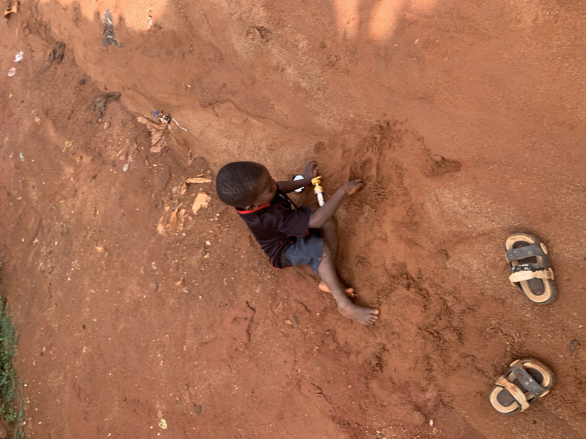 A child sits on bare, red earth in Ukwu-Abwa, Nigeria. He wears a dark shirt and blue shorts and reaches for a white and gold object. A pair of sandals lie nearby. Trash is scattered in the surrounding earth.
