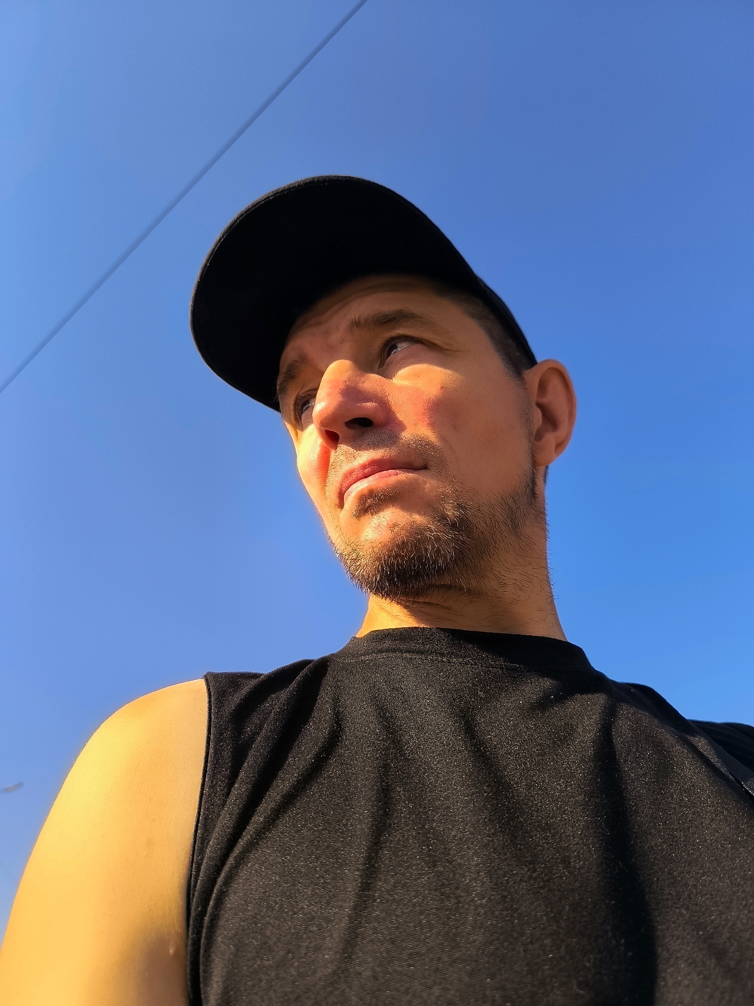 A low-angle, medium shot captures a man from the chest up, looking to the left against a clear blue sky. He wears a black sleeveless t-shirt and a black baseball cap. Sunlight illuminates the right side of his face, casting a warm glow on his skin and highlighting a light beard and stubble. The background is dominated by the expansive blue sky, with a thin, dark horizontal line, possibly a wire or cable, visible across the upper portion of the frame. The man's left shoulder and upper arm are also visible, bathed in bright sunlight. The image appears to be taken outdoors, possibly in a location with open sky. There is no indication of the specific location within Qarağandı city administration, Kazakhstan.