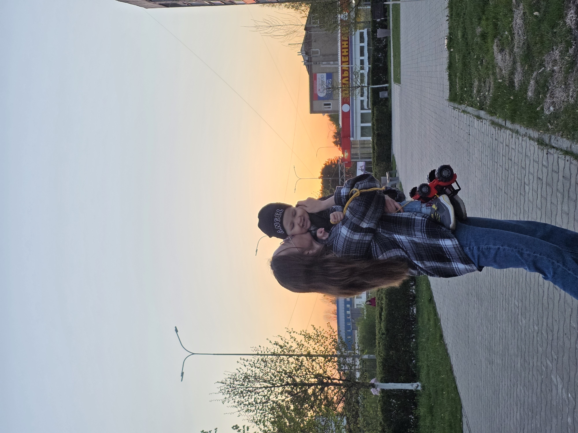 A vertical, full shot captures a mother and child outdoors, seemingly in Chaikovsky, Russia, given the building signage in the background. The mother, with long brown hair, wears a plaid shirt and blue jeans. She holds a young child, who is wearing a dark-colored hat with the word "ANGEL" on it and dark clothing. The child is turned towards the mother, with their faces close to each other.
The pair is standing on a paved walkway lined with green grass and bushes. To the right, a red toy truck rests on the ground. In the background, there is a building with signs in Russian Cyrillic. The sky is transitioning from light to dark, indicating either sunset or sunrise. Streetlights and trees are also visible in the background.