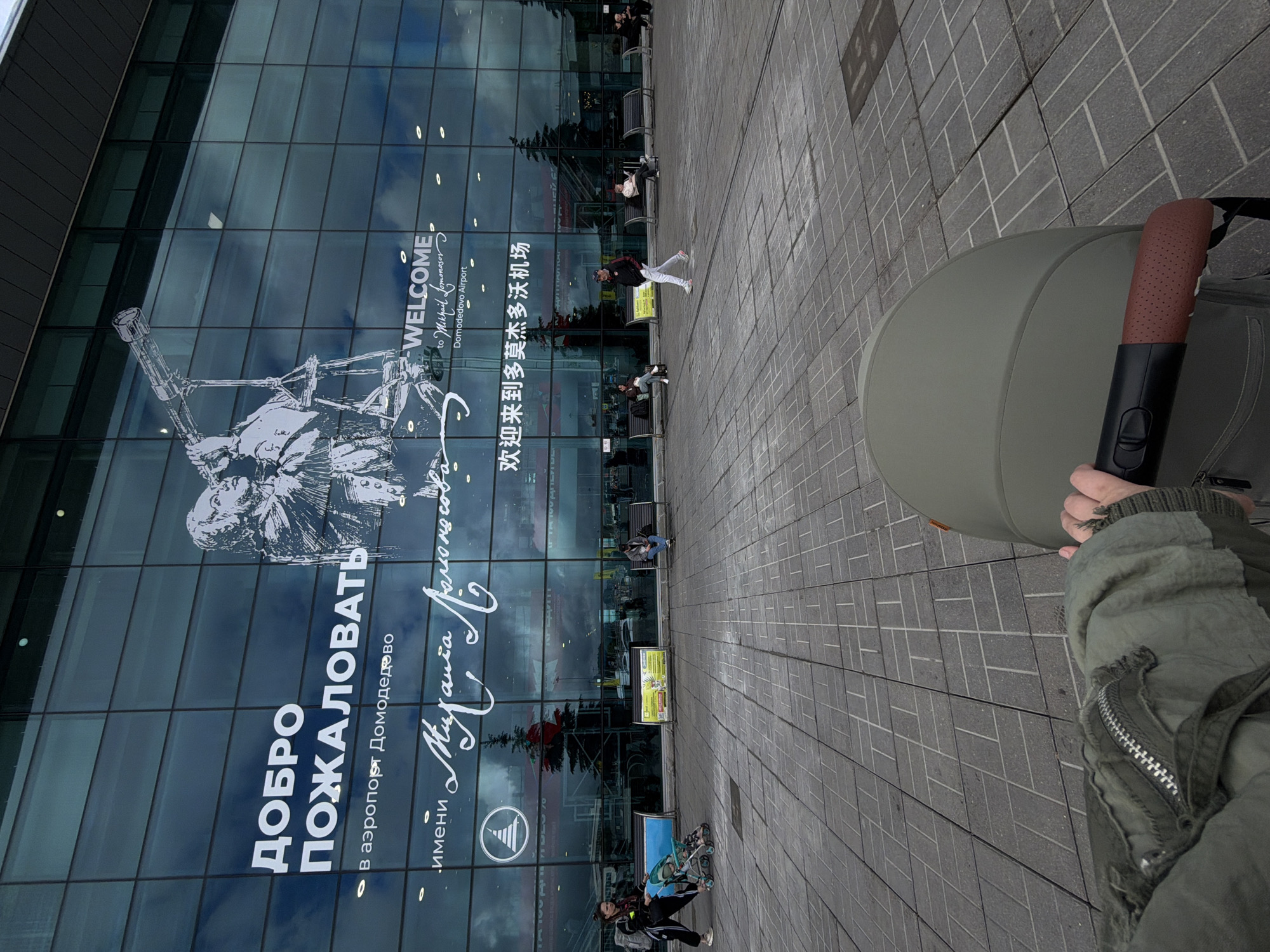The photograph was taken outside Domodedovo Airport, located in the city of Domodedovo, Russia. The building's glass facade displays a large welcome sign written in Russian, Chinese, and English, alongside an image of a telescope and a portrait of Mikhail Lomonosov. Outside the building, people can be seen walking and sitting on benches. The person who took the photo is pushing a light-green stroller, holding its brown handle. The stroller and the person's green jacket occupy the lower right corner of the photograph. The foreground is occupied by paved, gray-toned, squared-off stone blocks, suggesting a pedestrian walkway or plaza.