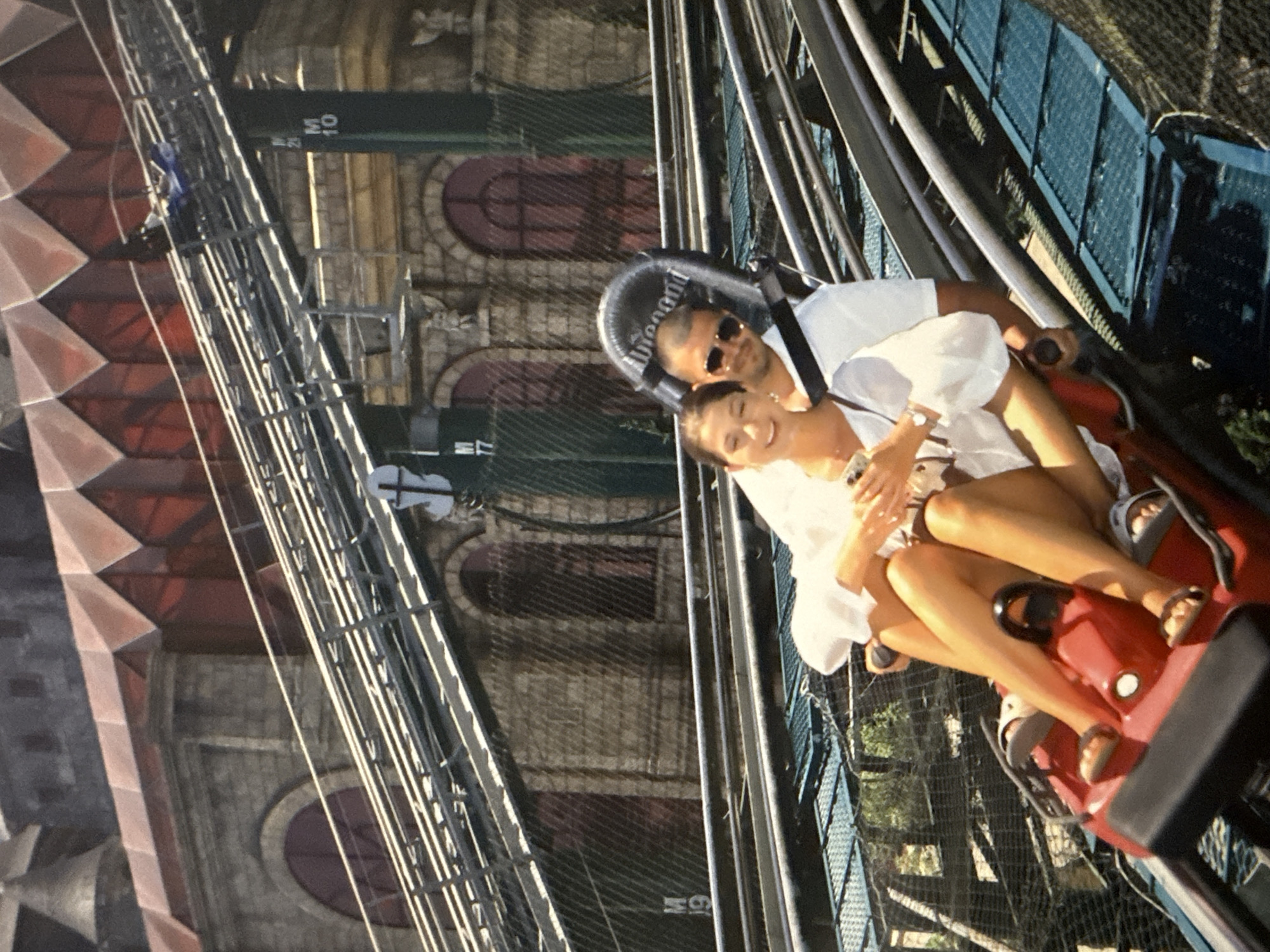 A couple rides on a coaster in Burgas, Bulgaria. The man in the back wears sunglasses and a white shirt, with a safety bar over his head. The woman in the front, also in white, holds a phone. The coaster's track and a building facade fill the background.