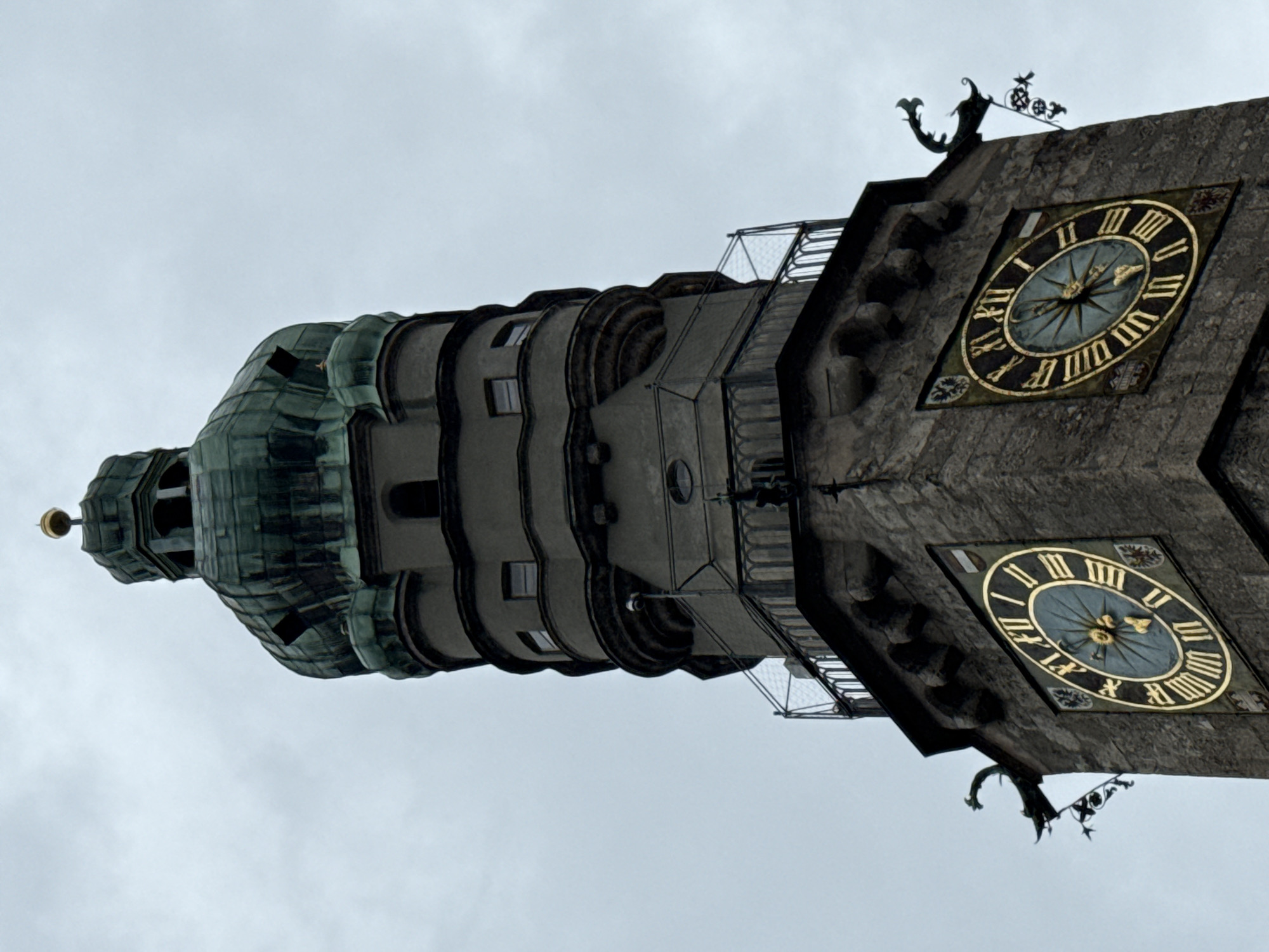 This is a low-angle, full-shot, eye-level, outdoor, vertical medium shot of the Rathausglockenturm, or City Hall Clock Tower, in Innsbruck, Austria. The tower, made of grey stone, has a green, octagonal, domed roof topped by a golden finial. Below the dome are several levels with small windows and decorative balconies. On the lower part of the tower are two large, round clock faces with Roman numerals and gold hands, set against a light blue background. The left clock shows approximately 4:05, and the right clock shows approximately 7:10. The clocks are adorned with small coats of arms on either side. Two decorative gargoyle-like figures in dark metal are mounted on the tower, one on each side of the clocks. The sky is overcast and grey, suggesting a cloudy day. There are no people or animals visible in the image.
