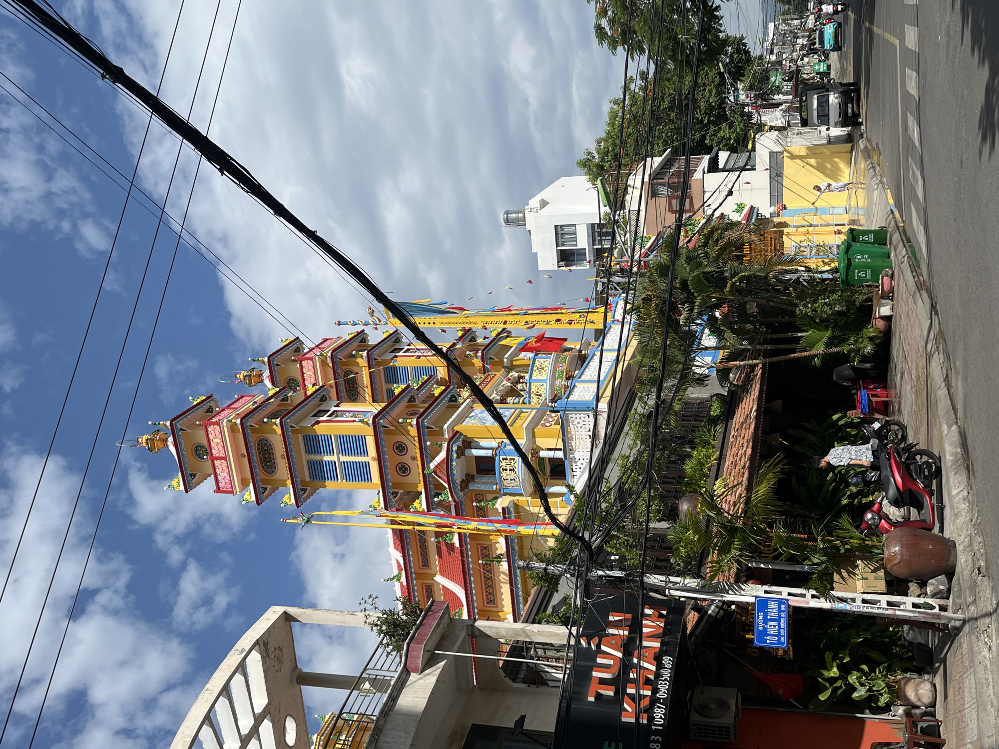The image captures a street scene in Nha Trang, Vietnam, dominated by a vibrant, multi-tiered Buddhist pagoda. The pagoda, painted in bright yellow, red, and blue hues, stands tall with ornate decorations and multiple levels. Power lines crisscross the sky, partially obscuring the structure. In the foreground, a sidewalk lines the street, with various businesses and signs visible. One sign reads "TUAN KHANH". A red scooter is parked on the sidewalk near a large ceramic pot, and a person in a patterned shirt stands beside it. Palm trees and other lush greenery are interspersed throughout the scene. The street itself is paved, with a crosswalk marked. A few vehicles, including a white van and a light blue car, are seen on the road in the distance. The sky is partly cloudy, with patches of blue visible.