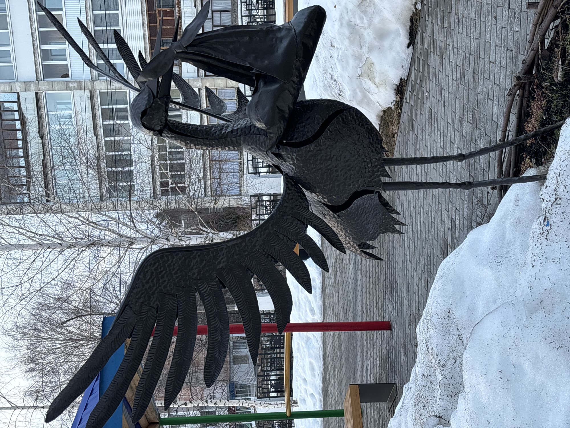 A large, dark-colored metallic sculpture of a stork is centrally positioned in a public outdoor area in the city of Chaikovsky, Russia. The stork has outstretched, textured wings and holds a dark, bundled object in its beak. The ground consists of gray rectangular paving stones with irregular patches of melting snow. To the left of the sculpture, parts of a playground are visible, including a blue roof section, a green vertical pole, a red vertical pole, and a light-colored wooden bench with a dark base. In the background, several bare deciduous trees with light bark are present, partially obscuring a multi-story, light-colored residential apartment building featuring numerous windows and enclosed balconies. A black metal fence is also visible in the mid-background. The overall scene suggests a late winter or early spring environment. No human figures are present.