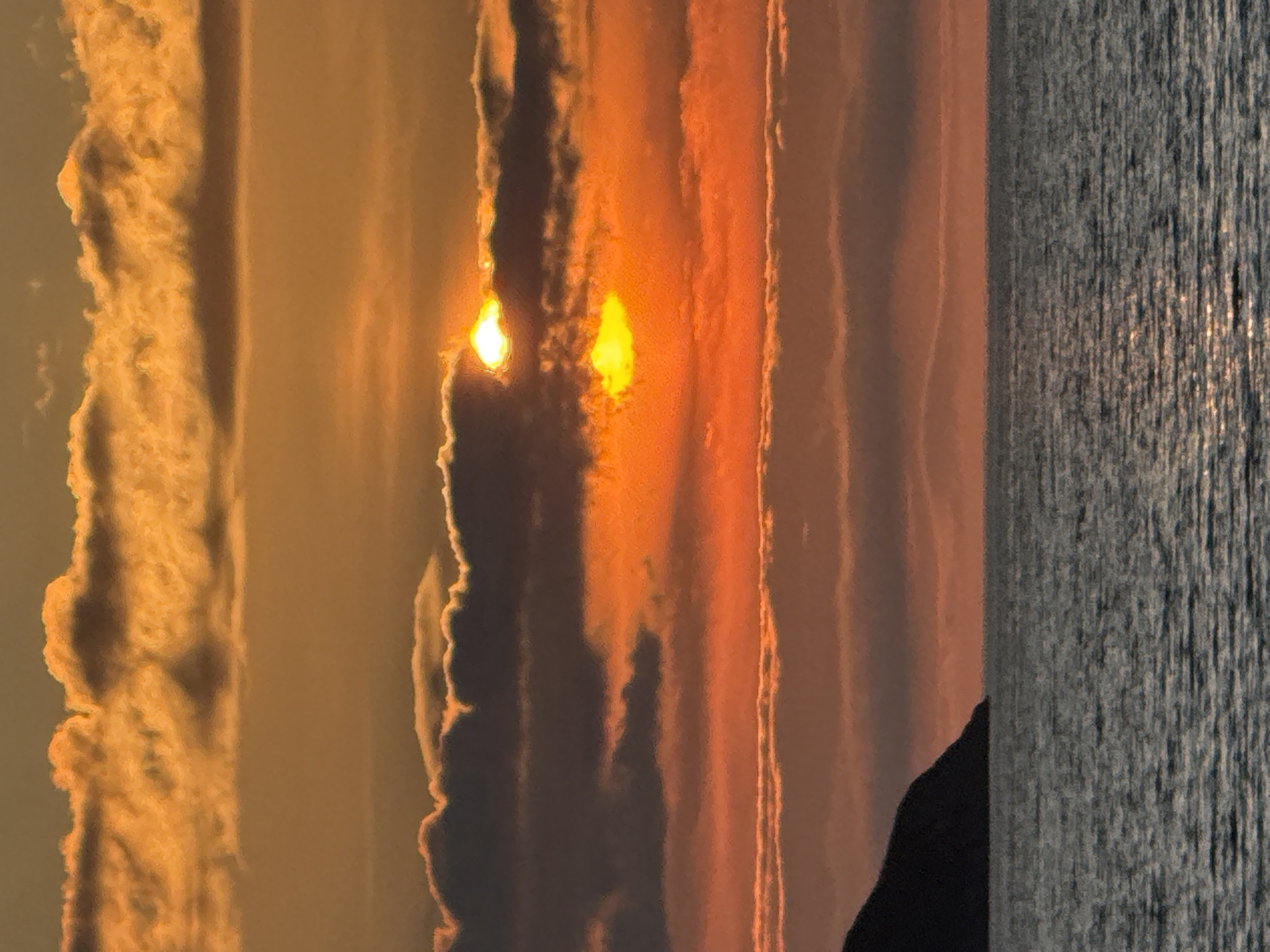 A dramatic sunset unfolds over the Barents Sea in Teriberka, Russia, casting the sky in hues of orange, yellow, and deep red. Clouds, illuminated from behind, create a textured and layered effect across the horizon. Partially obscured by these clouds, the sun appears as two bright, glowing orbs. The water below reflects the vibrant colors of the sky, with shimmering light dancing on its surface. To the left, the dark silhouette of a rocky shore or small island juts out into the sea, its form softened by the low light. The overall scene is one of natural beauty and the raw power of an Arctic sunset.