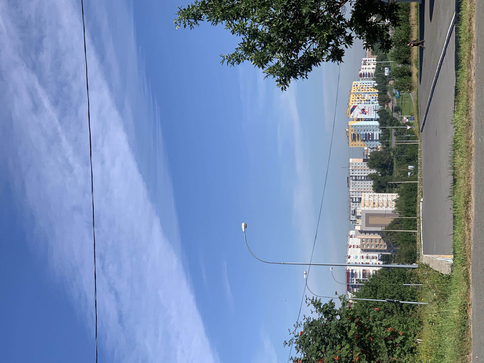 This is a daylight, eye-level, full outdoor shot of a city landscape in Prokopyevsk, Russia, taken from a slightly elevated perspective looking towards a row of apartment buildings. A dark wire runs horizontally across the bright blue sky, which is dotted with wispy white clouds. In the foreground on the left is a patch of green grass and a rowan tree with red berries. A street lamp is visible on the left, and another further back. A paved road with white markings curves from the lower right corner into the middle ground. A single person in dark clothing is walking on the road from right to left. Behind the foreground elements, a line of multi-story apartment buildings with various colored facades stretches across the horizon. Some buildings have balconies and visible windows. Green trees and vegetation are scattered between the buildings and in the surrounding landscape. A large billboard is visible on one of the colorful apartment buildings. The overall scene depicts an urban environment under a clear sky on a sunny day.