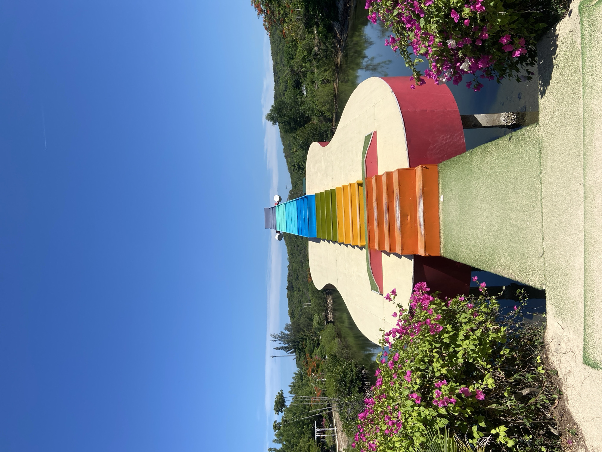 This is a photograph of a guitar-shaped bridge over a body of water in Hoa Thang, Vietnam. The bridge is painted in bright colors, with a rainbow-colored staircase leading up to the neck of the guitar. The body of water appears to be a lake or a slow-moving river, and it is surrounded by lush green vegetation and hills in the background. The sky is clear and blue. There are no people visible in the image. The bridge is a prominent structure in the landscape, and its unique design makes it a striking point of interest.