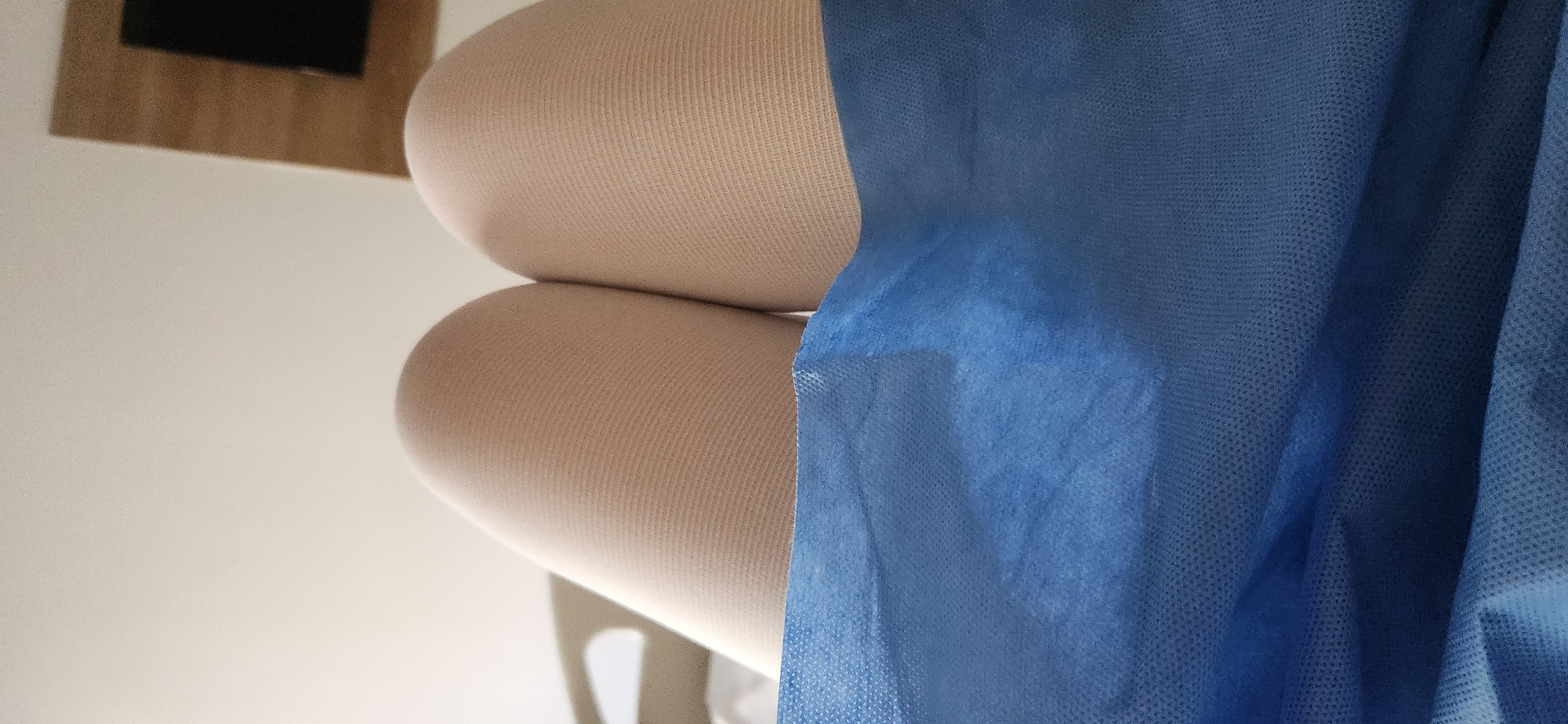 The image shows a close-up view of a person's legs, covered by compression stockings and a blue disposable medical gown. The legs are bent at the knee, and the compression stockings are a light tan color with a ribbed texture. The blue gown is draped over the lower portion of the legs, obscuring them from the waist down. The background is out of focus, but appears to be a room with a light-colored wall and a piece of wooden furniture. There are no discernible features to indicate the location in Beşiktaş, Turkey.