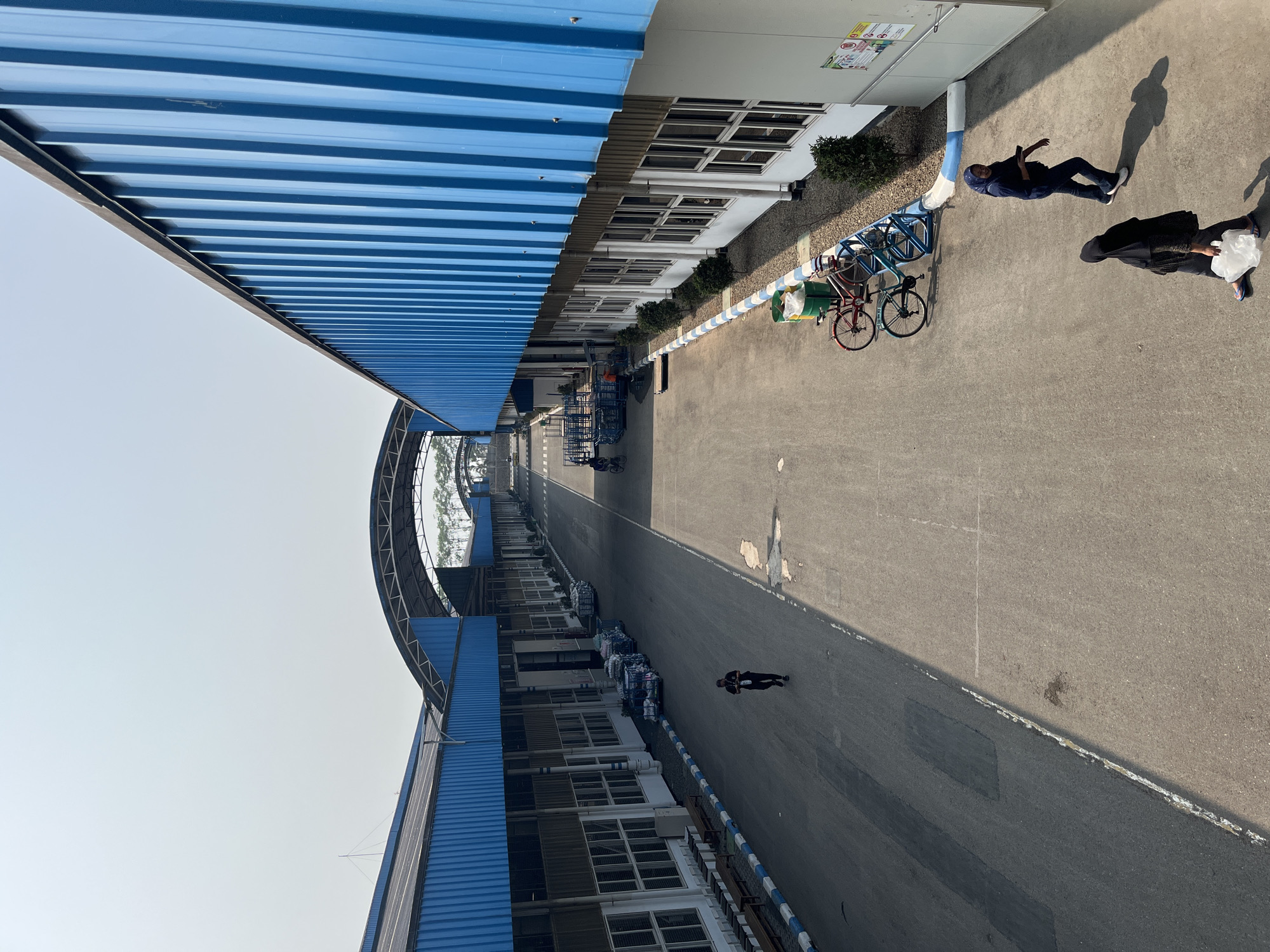 The image shows an outdoor paved area with buildings on either side, a perspective taken from an elevated position looking down an avenue. The buildings feature blue corrugated metal roofs supported by a structure that creates covered walkways or loading docks. These buildings have rows of windows. The ground is a light gray asphalt with some darker patches and white markings along the edges.
On the left side of the avenue, there are benches and some industrial carts. A lone person in dark clothing is walking away from the camera down the middle of the avenue.
On the right side of the avenue, near the foreground, two women are walking. One woman, wearing a dark hijab and a patterned top, is carrying a white plastic bag. The other woman, wearing a blue hijab and dark clothing, is looking towards the camera and holding a phone. Behind them are two bicycles parked at a blue rack, one red and one blue. Further down the right side, there are small, manicured bushes planted along the building.
In the distance, the avenue continues, with more industrial carts and some trees visible. The sky is clear and light blue.
The provided context states the location is the city of Pelang, Indonesia. This scene appears to be a factory or industrial complex within that city.