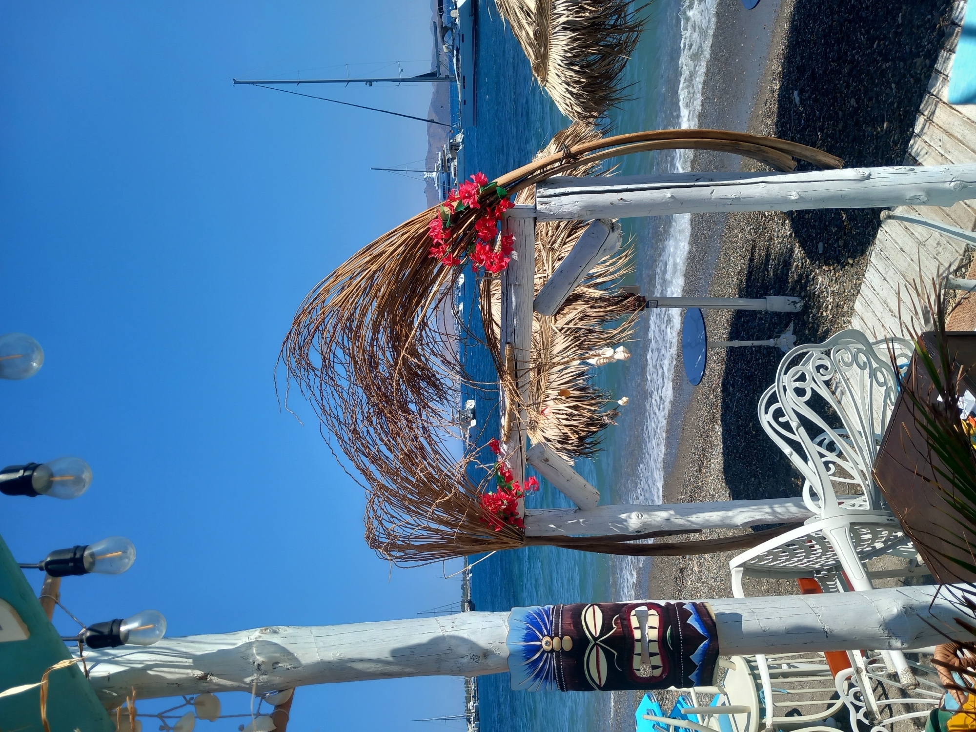 This image depicts a sunny day at a beachfront establishment in the city of Kos, Greece. A white wooden archway is adorned with dried palm fronds and a cluster of bright red flowers. In front of the archway, a tiki mask is mounted on a weathered white wooden post. Several white chairs and tables are visible on a wooden deck in the foreground. Beyond the deck, a pebble beach slopes gently towards the turquoise waters of the Aegean Sea. Several sailboats and yachts are anchored in the bay, and a mountainous coastline is visible in the distance. The overall atmosphere is serene and inviting, suggesting a perfect spot for relaxation and enjoyment of the Mediterranean coast.