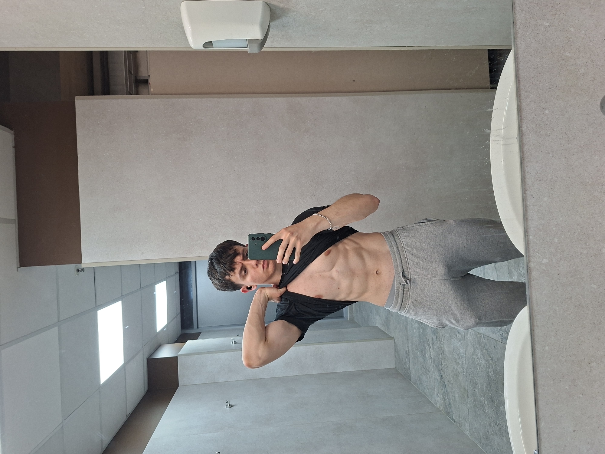 A young man is posing for a mirror selfie in a bathroom. He is wearing grey sweatpants and a black t-shirt, which he is pulling up to expose his torso. The bathroom has white tiled walls and floor, and a white sink is visible. The location is potentially in Plovdiv, Bulgaria, but this cannot be confirmed from the image.