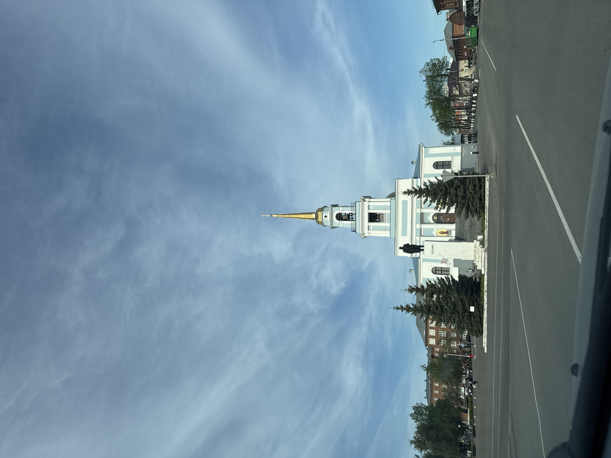 The image shows a view of the city of Votkinsk, Russia. A church with a golden spire is prominently featured in the foreground. Next to the church is a statue of Lenin. A road runs alongside the church. Buildings are visible in the background. The sky is blue with wispy clouds.
