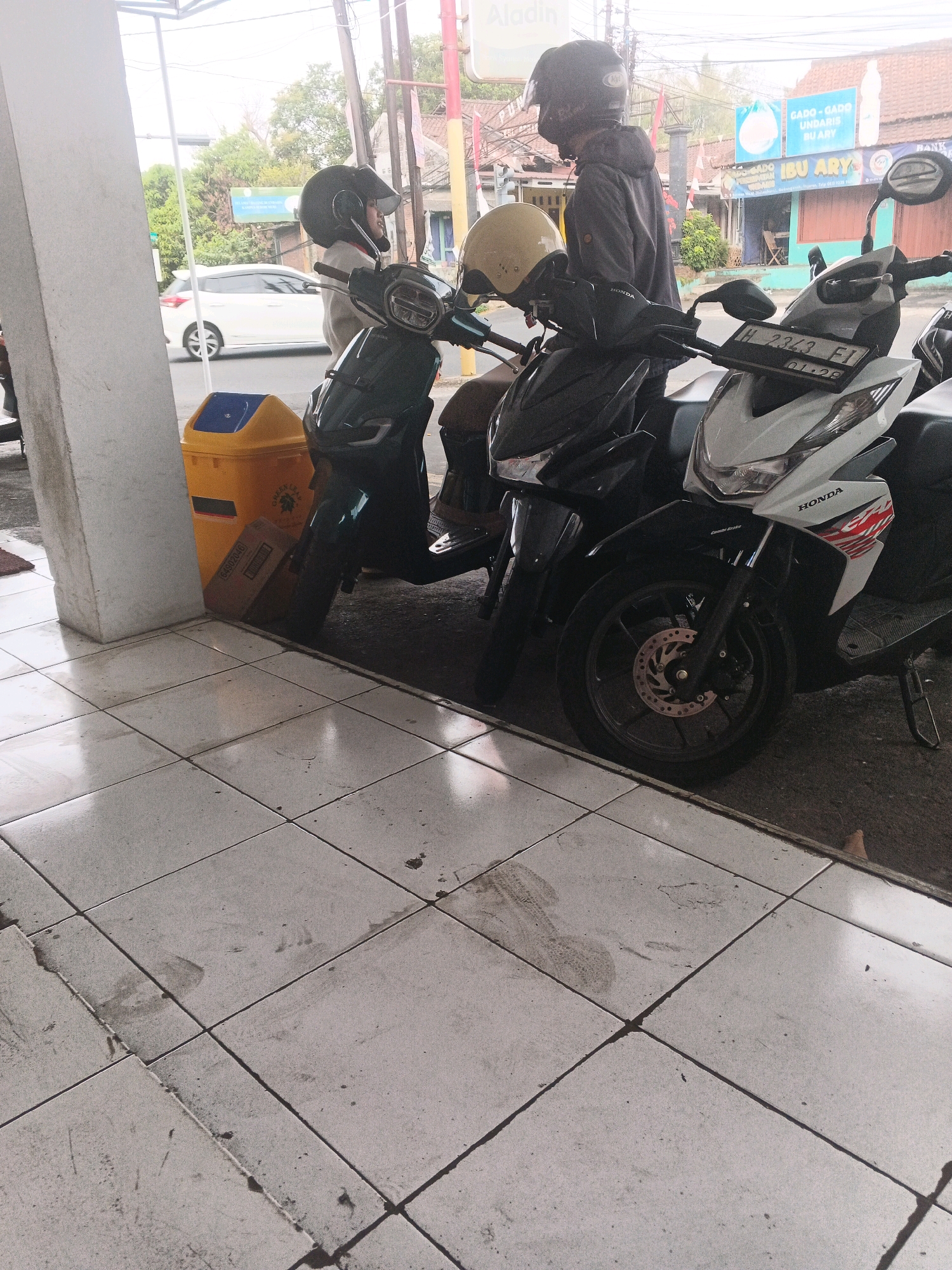 Two individuals wearing helmets are standing near two parked motorcycles on a tiled walkway. A yellow and blue trash can is visible to the left of the motorcycles. In the background, a white car is driving on a street, and buildings with signs are present. One sign reads "GADO-GADO IBUL ARY". The scene appears to be in a commercial or public area in Sidomulyo, Indonesia. The lighting suggests it is daytime.
