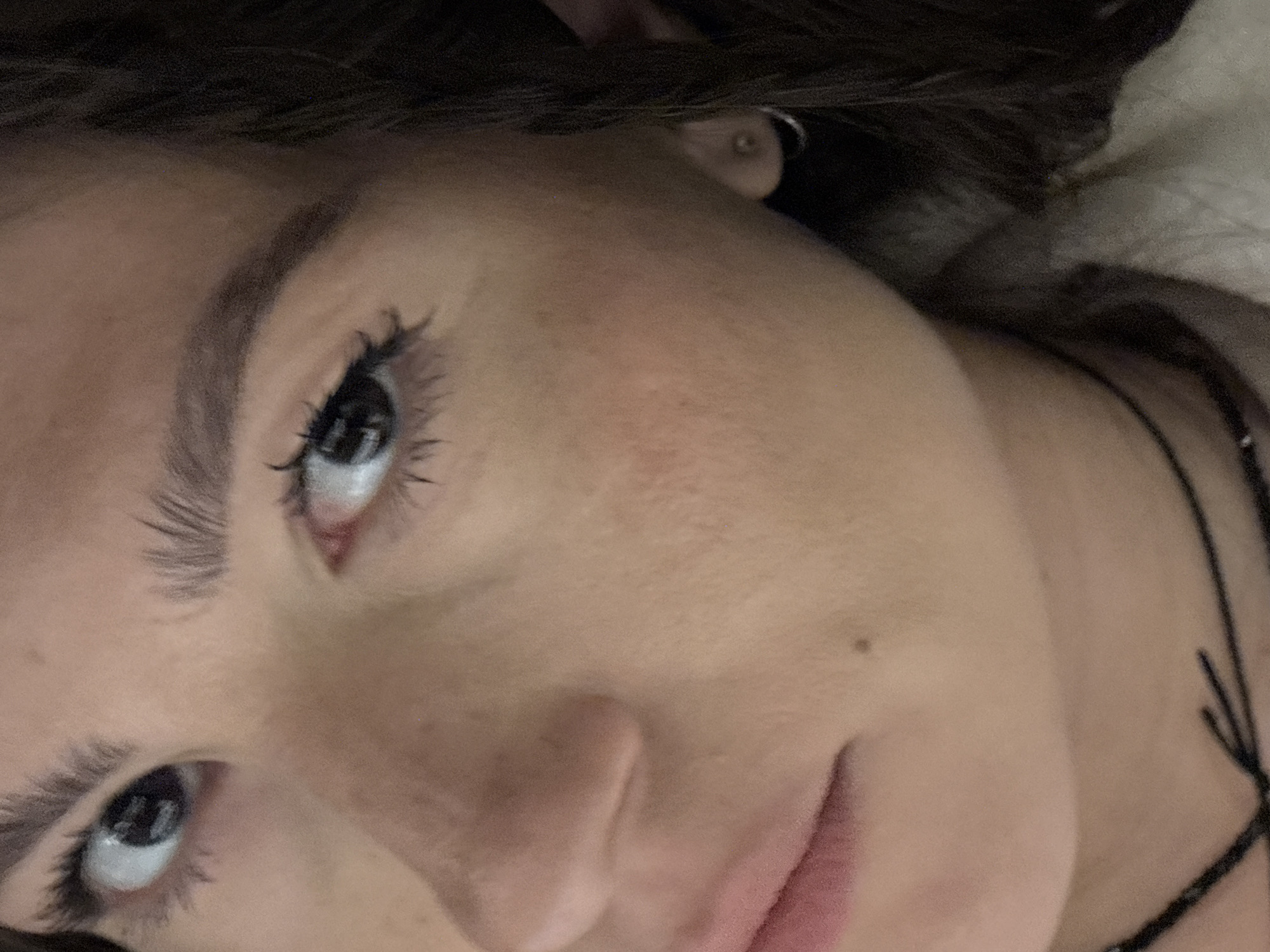 A close-up, angled view of a woman's face. She has dark hair and eyebrows, fair skin, and dark eyelashes. Her eyes are open and looking upward. A small mole is visible on her cheek. A necklace with black strings is around her neck. The background is blurred and appears to be a light color, possibly a pillow or blanket. The image is not geographically contextualized in Шемордан, Russia or any other location.