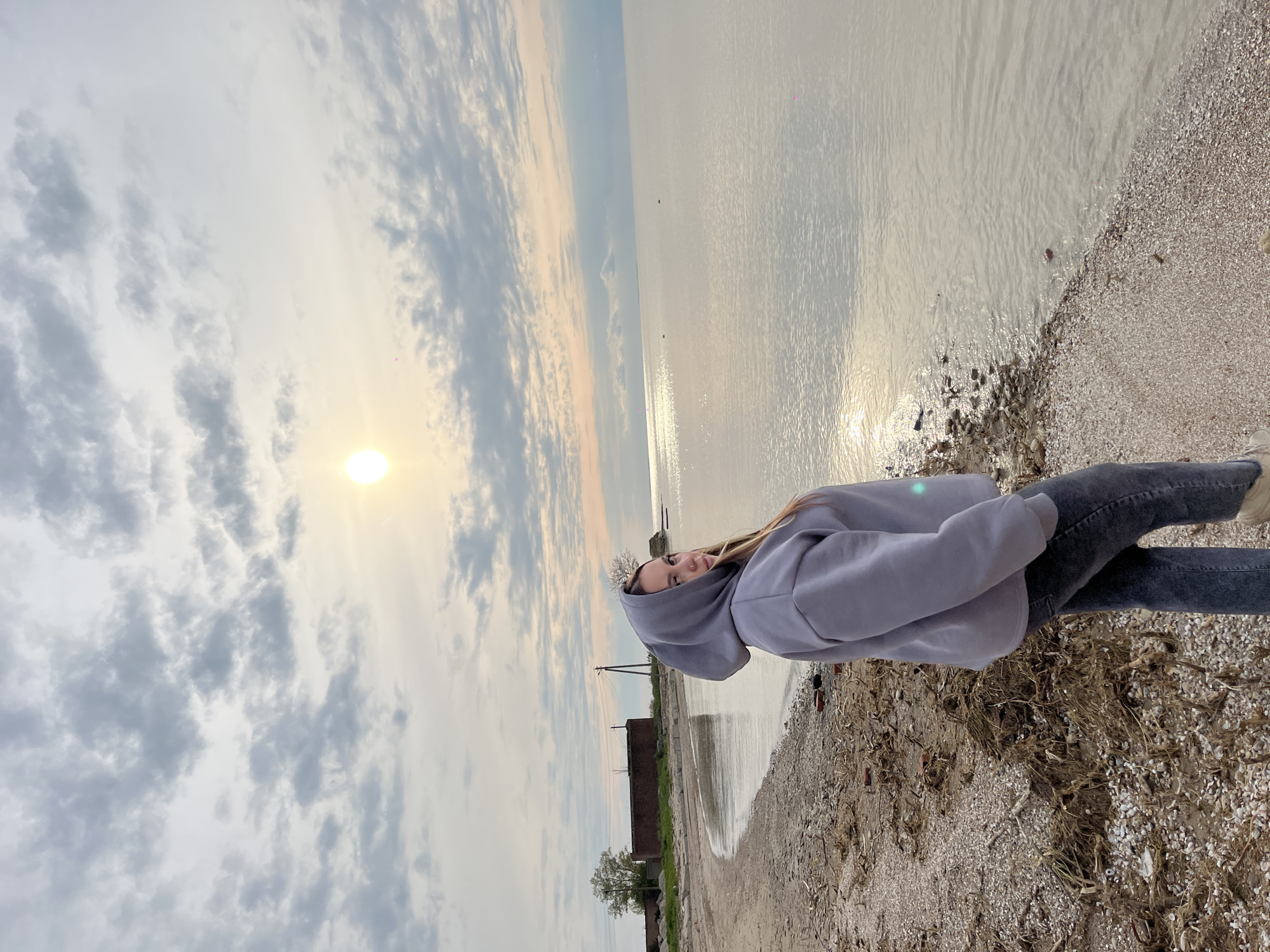 A young woman stands sideways on the pebbly shoreline of Port-Katon, Russia, facing the camera. She is wearing a light grey hooded sweatshirt and dark trousers. Her long hair flows over her shoulders. The image is rotated 90 degrees clockwise, making her appear to be lying on the ground next to the water.
The scene depicts a calm body of water, likely a bay or large lake, stretching to the horizon under a partly cloudy sky. The sun is low on the horizon, casting a bright glare on the water and through the clouds, indicating either sunrise or sunset. The shoreline is composed of small stones, shells, and significant amounts of dried seaweed and other natural debris.
In the background, along the distant shore, some low-lying structures resembling old buildings or ruins are visible, along with a utility pole and a sparse tree. No specific activity beyond posing for a photograph is evident. The overall atmosphere is serene, with the soft light of the sun reflecting off the water. There is no visible text in the image.