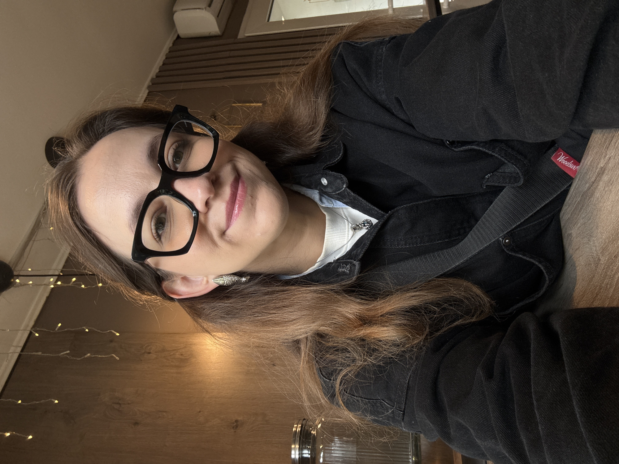 A medium shot shows a woman with long, brown hair, wearing black-rimmed glasses, a white undershirt, and a black denim jacket. She is looking at the camera and smiling. She has on silver fish-shaped earrings and a silver pendant necklace. The background is a wooden wall with string lights and a glass jar. This scene appears to be indoors, possibly in a cafe or restaurant. No specific location in Chaikovsky, Russia can be identified from the image.