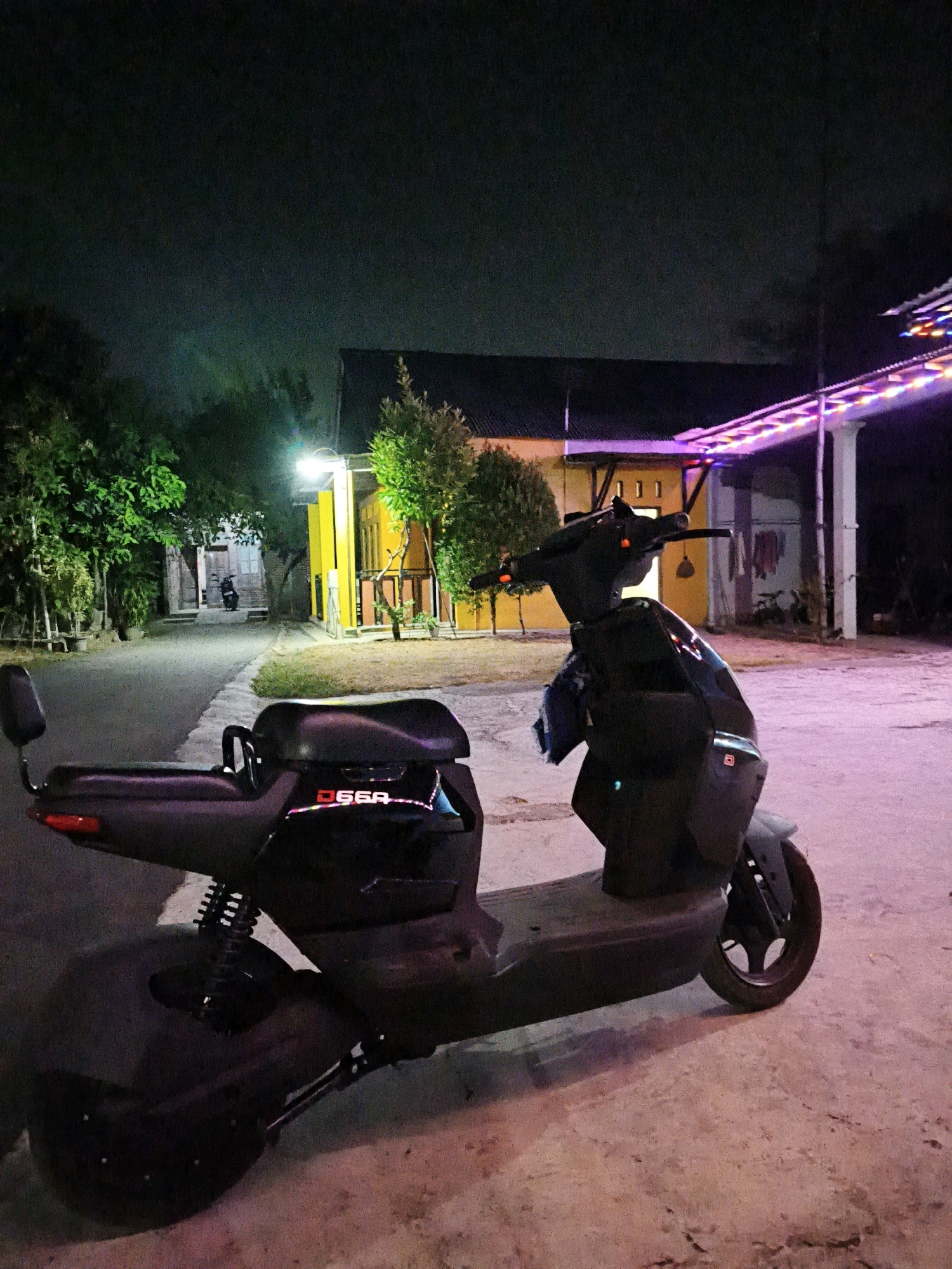 A dark, unbranded electric scooter is parked on a paved surface at night in front of a yellow building. The scooter is mostly black, with a seat and handlebars visible. A blue cloth is hanging from the handlebars. The building has a dark roof and several windows. There are trees and a road in the background. Lights from the building and a string of colored lights on an overhang illuminate the scene. The sky is dark and appears cloudy. The location is identified as Pengkok, Indonesia.