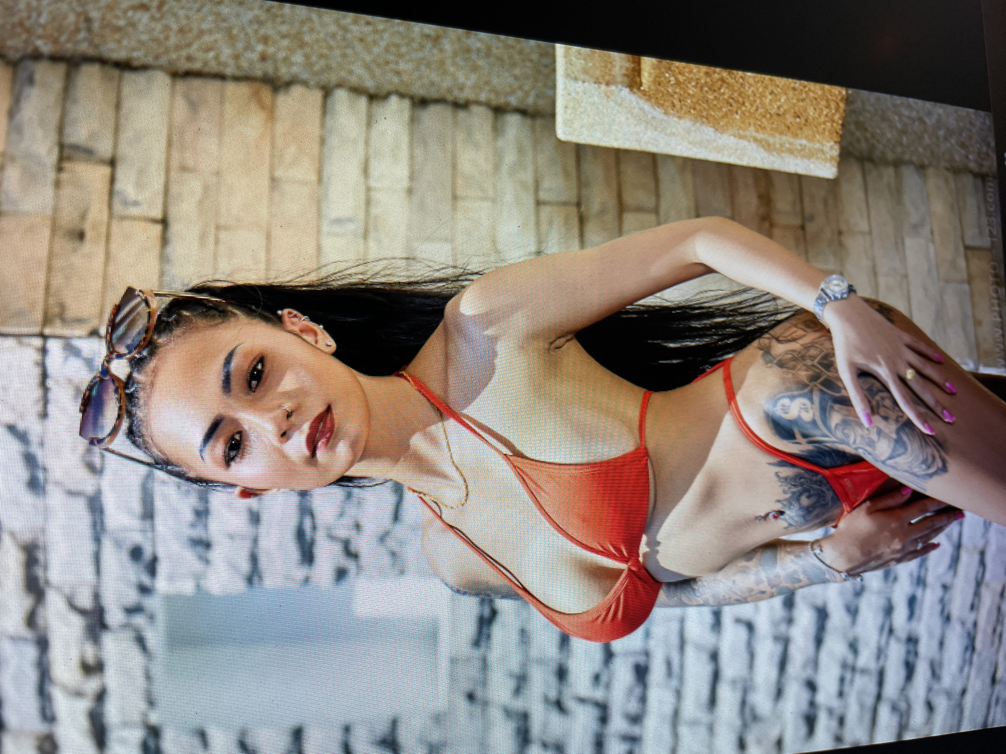 A woman is lying beside a swimming pool at Atmosphere Resorts & Spa in Bacong, Philippines. She is wearing a red bikini and sunglasses. The background includes stone flooring and other features of the resort. The woman has dark hair and multiple tattoos. She is wearing a wristwatch and a necklace. Her fingernails are painted pink.