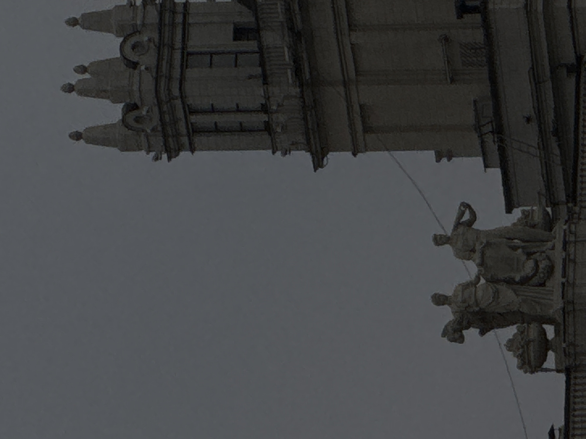 The image shows a section of a building exterior, specifically architectural details of what appears to be a tower or spire with statues. The architectural style suggests the image is from the city of Moscow, Russia. The building features ornate details including statues of human figures and what appears to be detailed stonework. The background is a grayish sky.