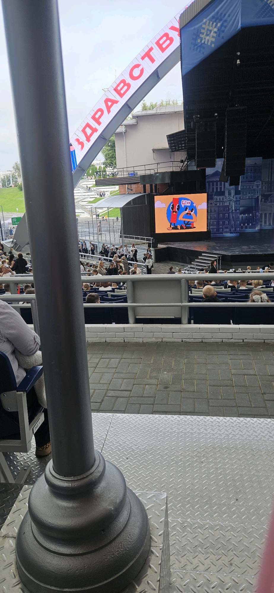 The image shows an outdoor venue with a large stage and seating area, likely for an event or performance. A prominent banner with large red Cyrillic lettering, reading "ЗДРАВСТВУЙ" (Zdravstvuy, meaning "Hello" or "Greetings" in Russian and Belarusian), is visible spanning across the top of the scene, angled upwards. To the right of the stage, a large LED screen displays a cartoonish graphic of two figures, one red and one blue, interacting. Rows of blue seats are visible in the foreground and midground, with people seated in them. A large grey pole or pillar is positioned in the left foreground, partially obscuring the view of the audience and the stage. The ground in the foreground and midground is paved with interlocking gray bricks and sections of diamond plate metal flooring. The background shows buildings and greenery, suggesting an urban setting. The overall atmosphere appears to be that of a public gathering or festival. The presence of Cyrillic script and the architectural style are consistent with locations in Belarus, and specifically Vitsebsk, which is known for hosting large cultural events like the "Slavianski Bazaar" festival, often featuring outdoor stages and elaborate decorations.