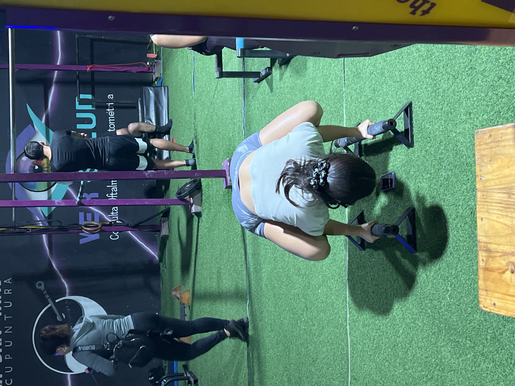 The scene depicts a fitness center in Arica, Chile. A woman in a white shirt and blue shorts is performing a rowing exercise with parallel handles on a green artificial turf floor. To the left of her, another woman wearing a grey jacket and black leggings is harnessed to a bungee fitness system. In the background, a man in black workout attire is lying on a padded surface. Behind the bungee fitness station is a wall with an advertisement featuring the words "acupuntura" and "oftalmológico".