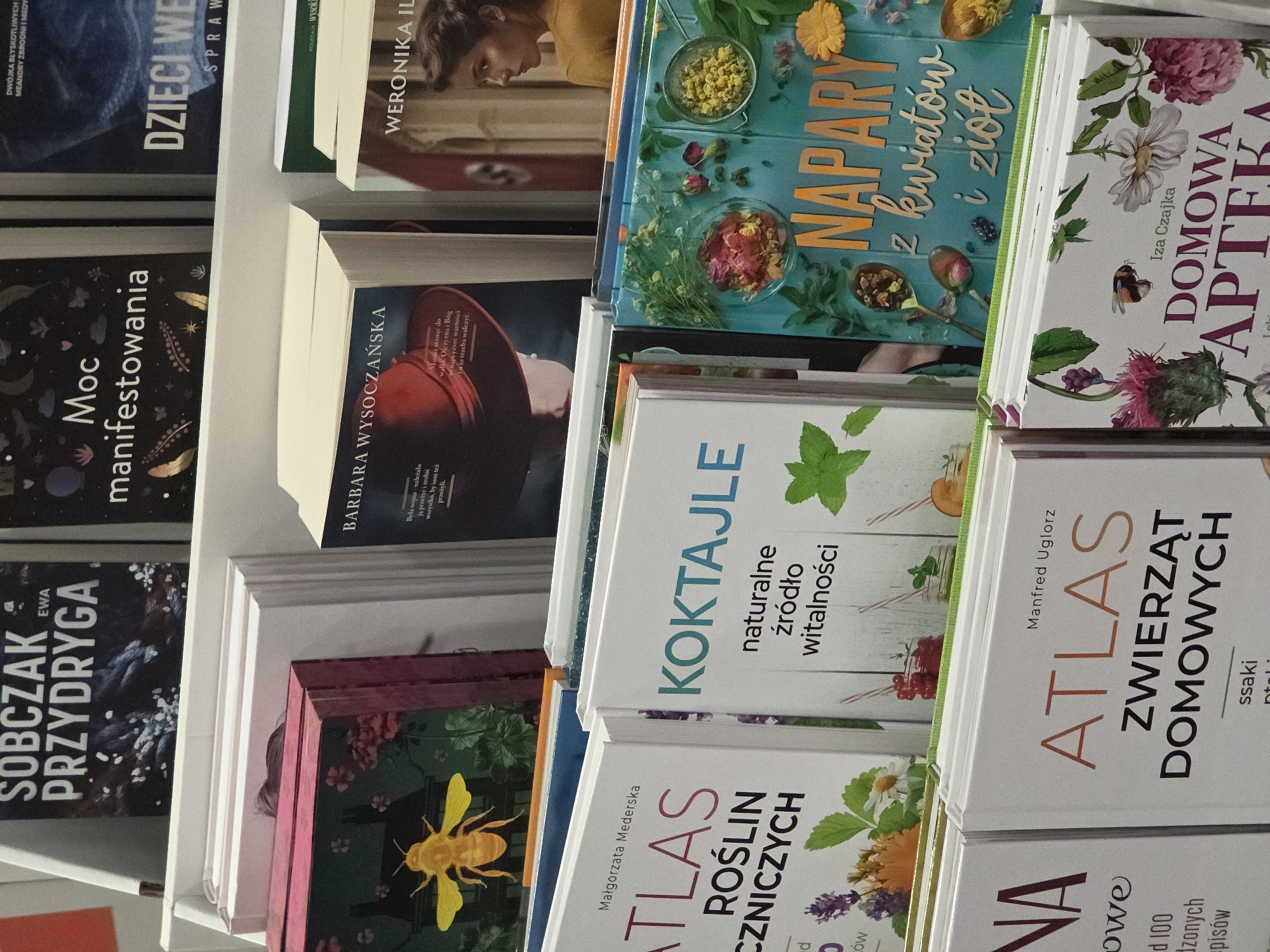 This image shows a shelf of books in a bookstore. The books are stacked on top of each other, and their spines are visible. The book titles and authors are in Polish. The books appear to be on topics such as herbalism, cocktails, and domestic animals.