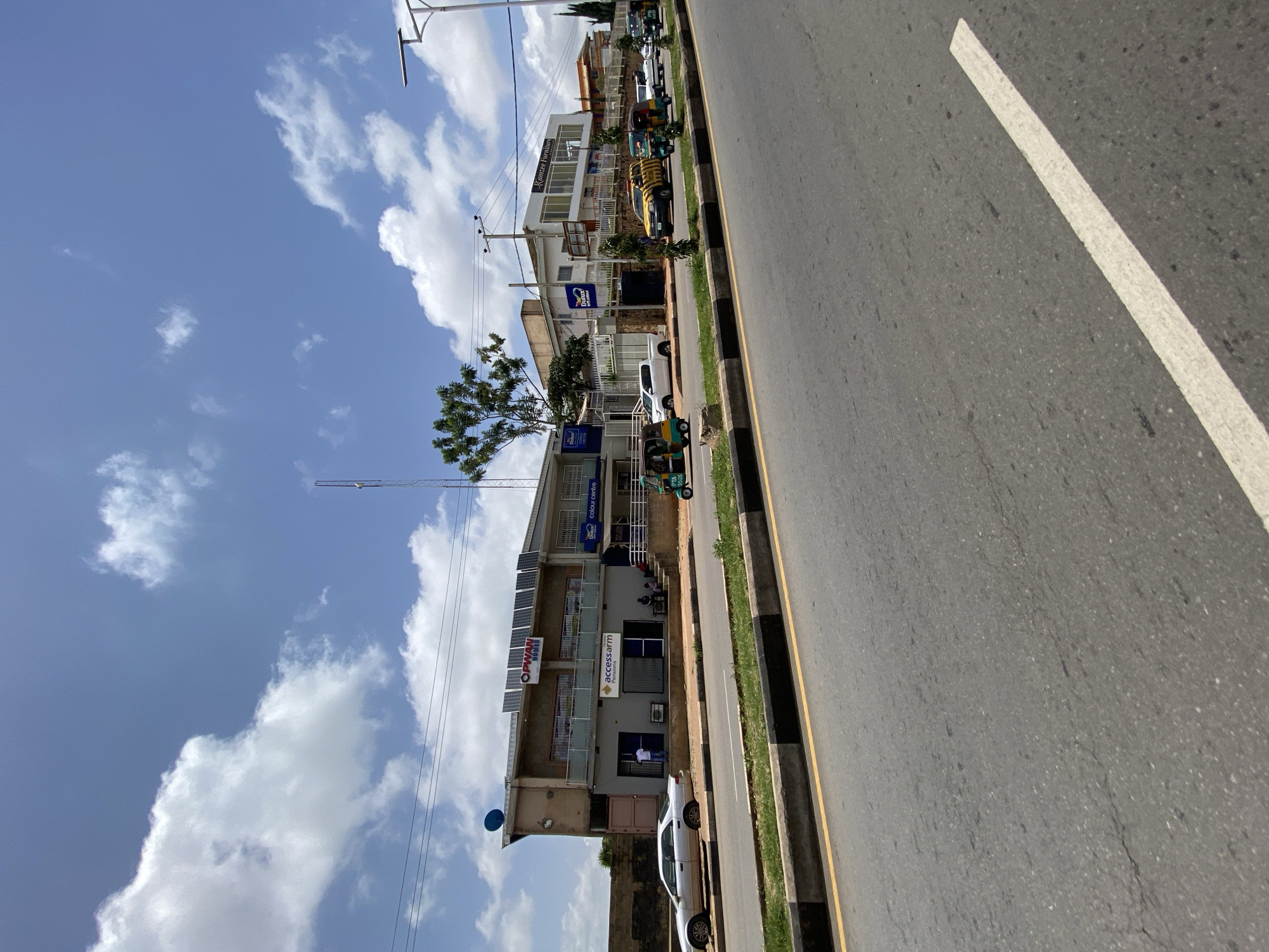 The image, taken from a low-angle perspective on a street in Jos, Nigeria, showcases a commercial building complex under a partly cloudy blue sky. The building features a modern design with a grey facade on the lower level and a lighter-colored upper story. Solar panels are installed on the roof of the lower section, and a satellite dish is visible on the left side of the building. Signs indicate businesses such as "OPWAN HOMES," "accessarm Pensions," "Dulux Colour Centre," and "Ramzee Furniture."
Several vehicles are present on the road and in front of the buildings, including a silver sedan, a white pickup truck, several three-wheeled auto-rickshaws (known locally as Keke Napep), and a yellow and black striped vehicle. People are seen standing and sitting in front of the businesses, and one person is standing by the silver sedan. The street is paved, with distinct yellow and black striped curbing along the side. A street lamp is visible on the right side of the image, and a telecommunications tower stands in the distance. The overall scene depicts a typical day in a commercial area of Jos, Nigeria.