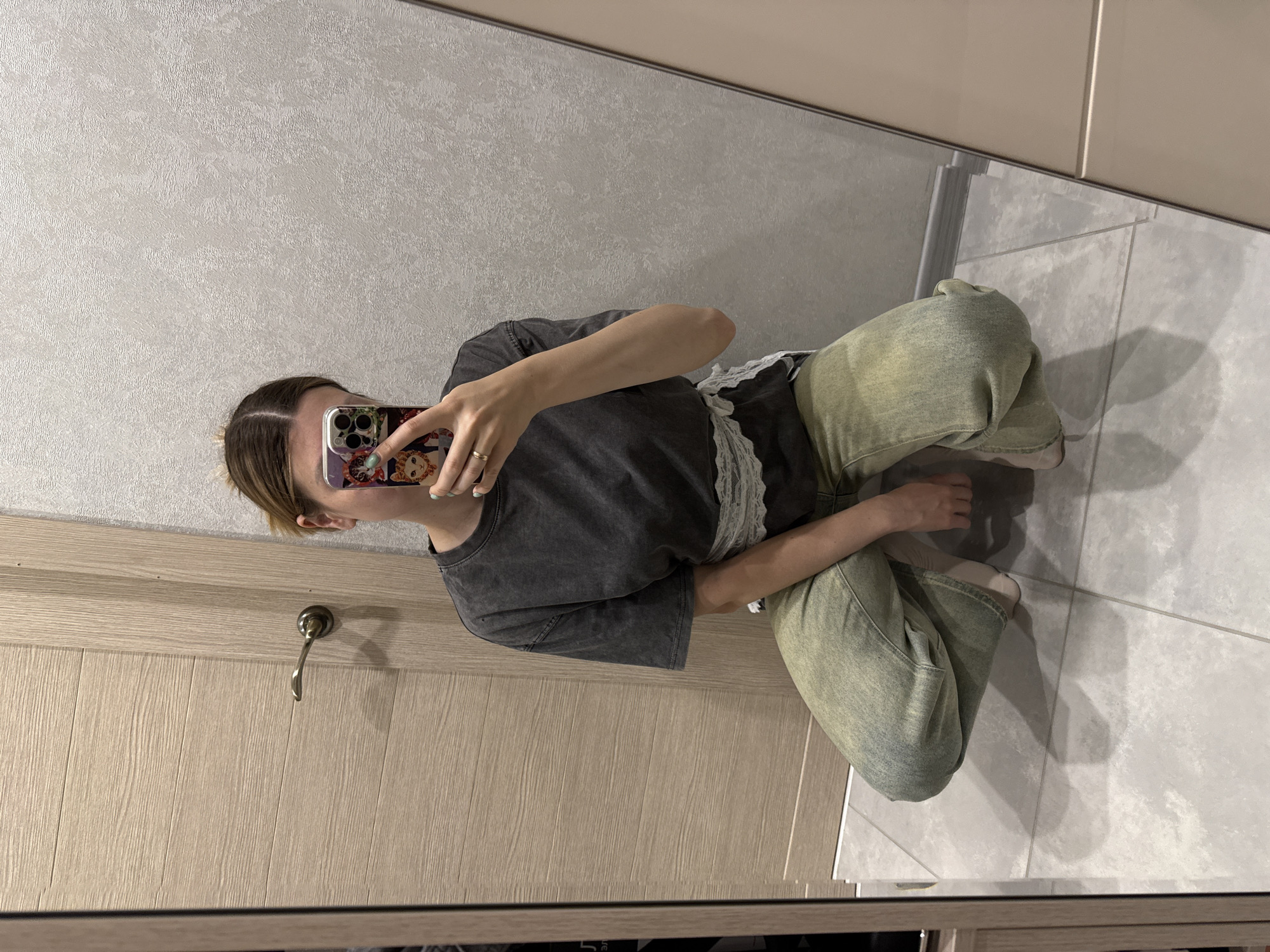 A mirrored image depicts a person squatting in a room with light grey textured wallpaper, beige wood-paneled doors with a silver handle, and light grey tiled flooring. The person is holding a phone with a decorative case, obscuring their face. They are wearing a grey t-shirt with a white lace trim and faded green pants. The reflection indicates the scene is indoors, with a cabinet visible near the ceiling. The city or country cannot be determined from the image.