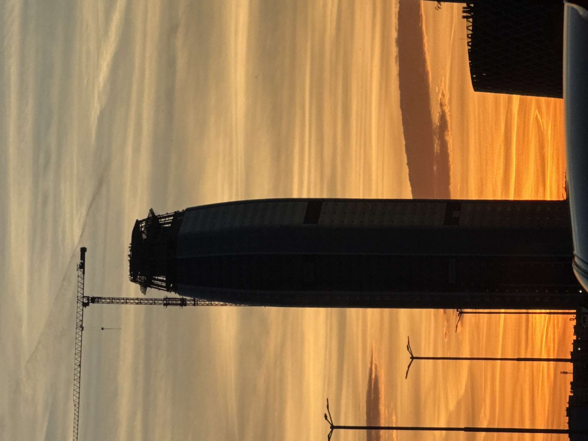 The image shows the silhouette of a building under construction against a striated sunset sky. A tall construction crane is visible to the left of the building. The building is elongated and curved on top, with a darker facade and areas appearing to be incomplete. In the foreground, there are lamp posts. Another building is visible to the right, as well as an additional white-colored element. The location is likely Sofia, Bulgaria due to the specific architectural style of the building under construction.